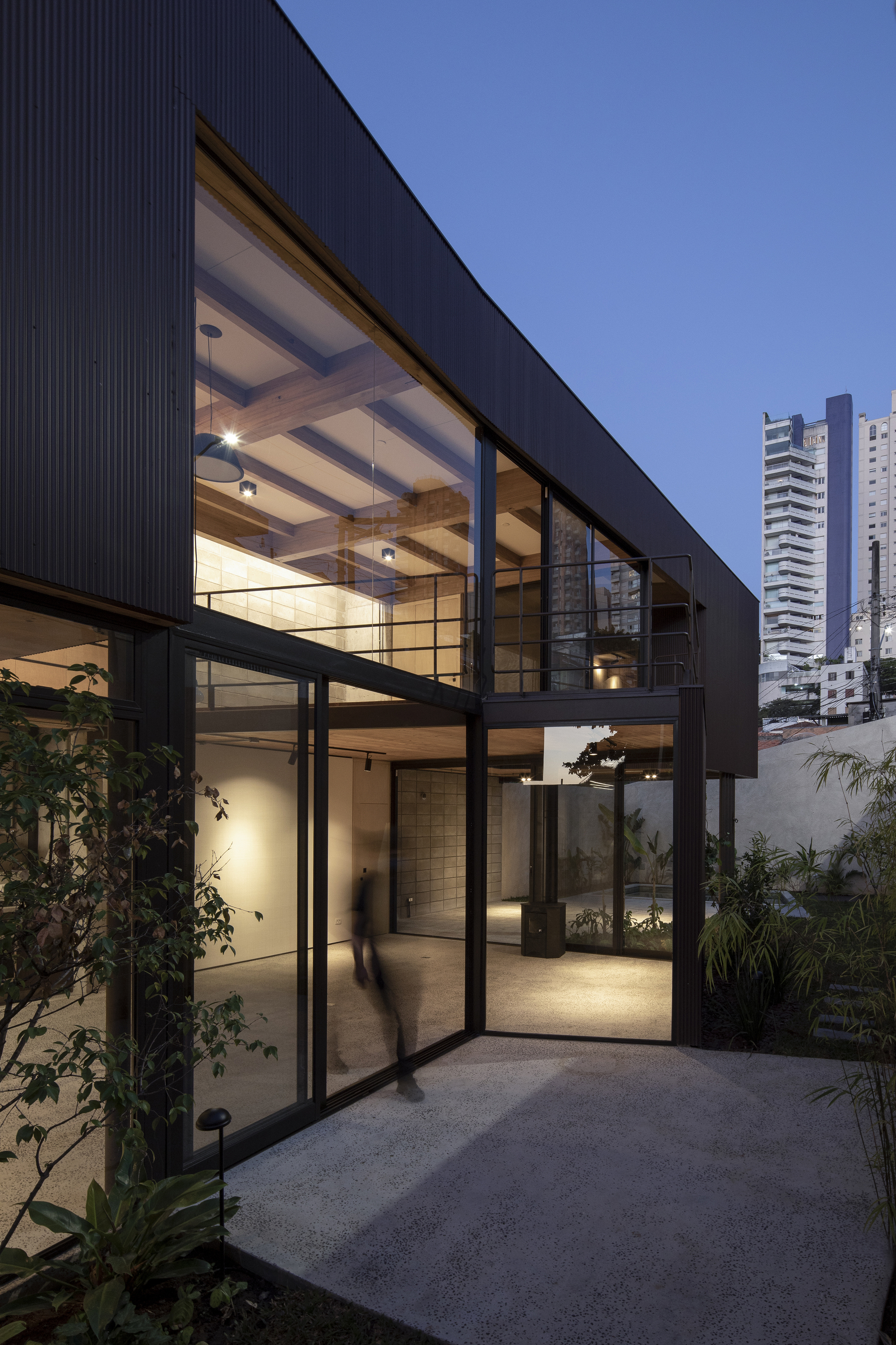 Glazed facade with black steel frames and corrugated metal cladding illuminated at dusk with tower beyond