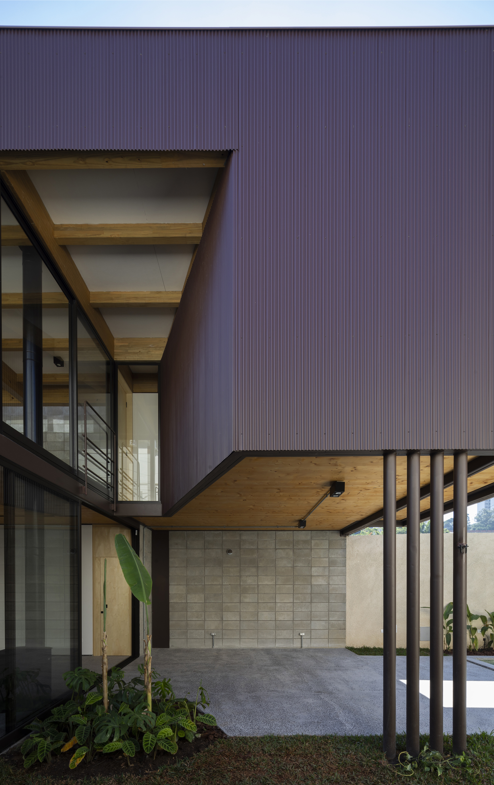 Covered entry courtyard with corrugated metal walls, timber soffit, concrete tile wall and planted border at dusk