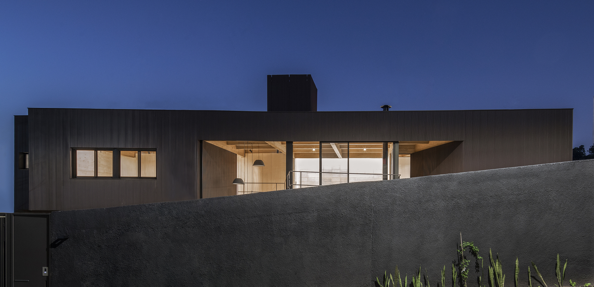 Dark corrugated metal facade with illuminated interior visible through large glazed openings at dusk