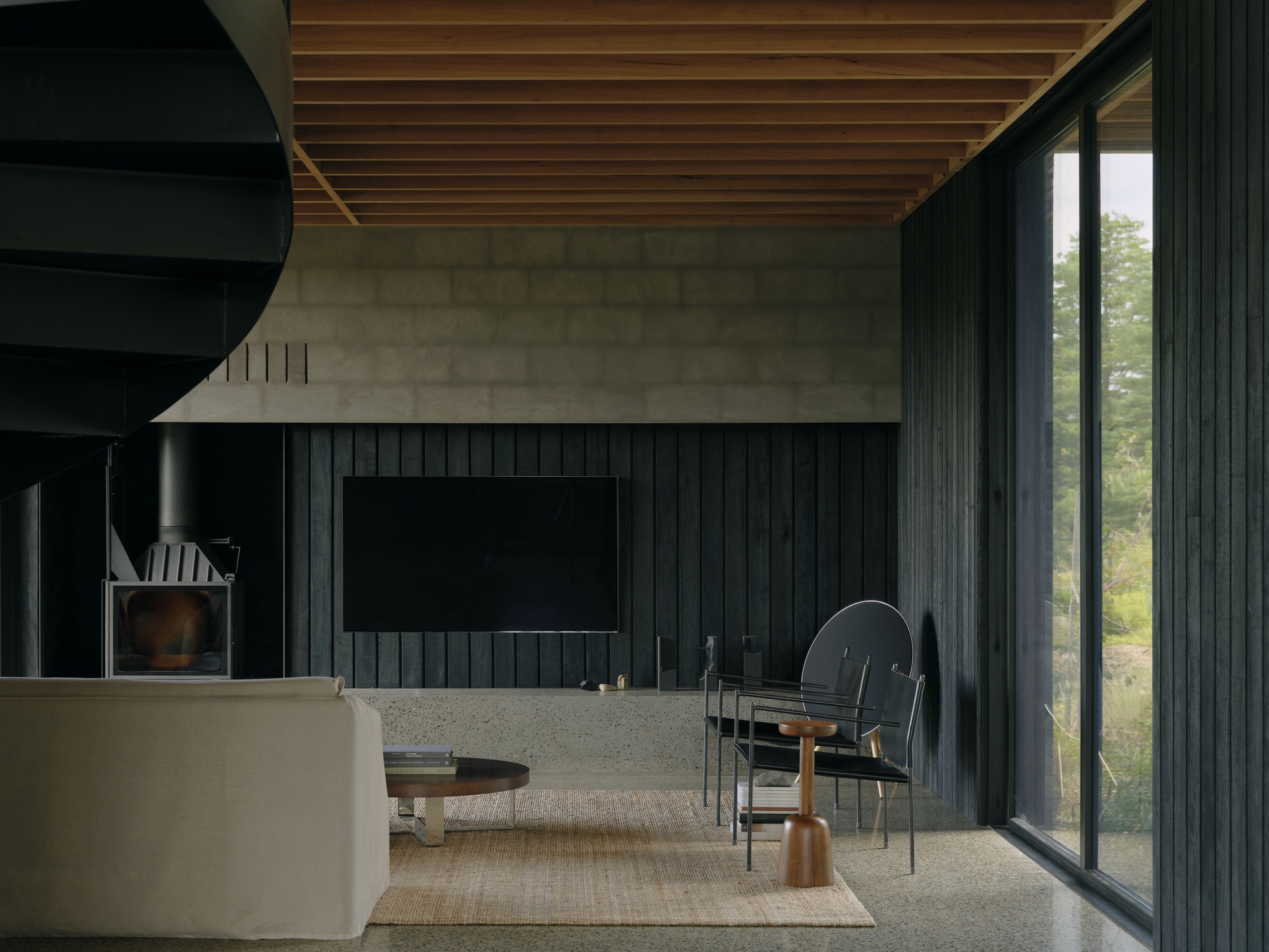 Living room with exposed timber ceiling beams, blackened timber cladding, and floor-to-ceiling glass overlooking landscape