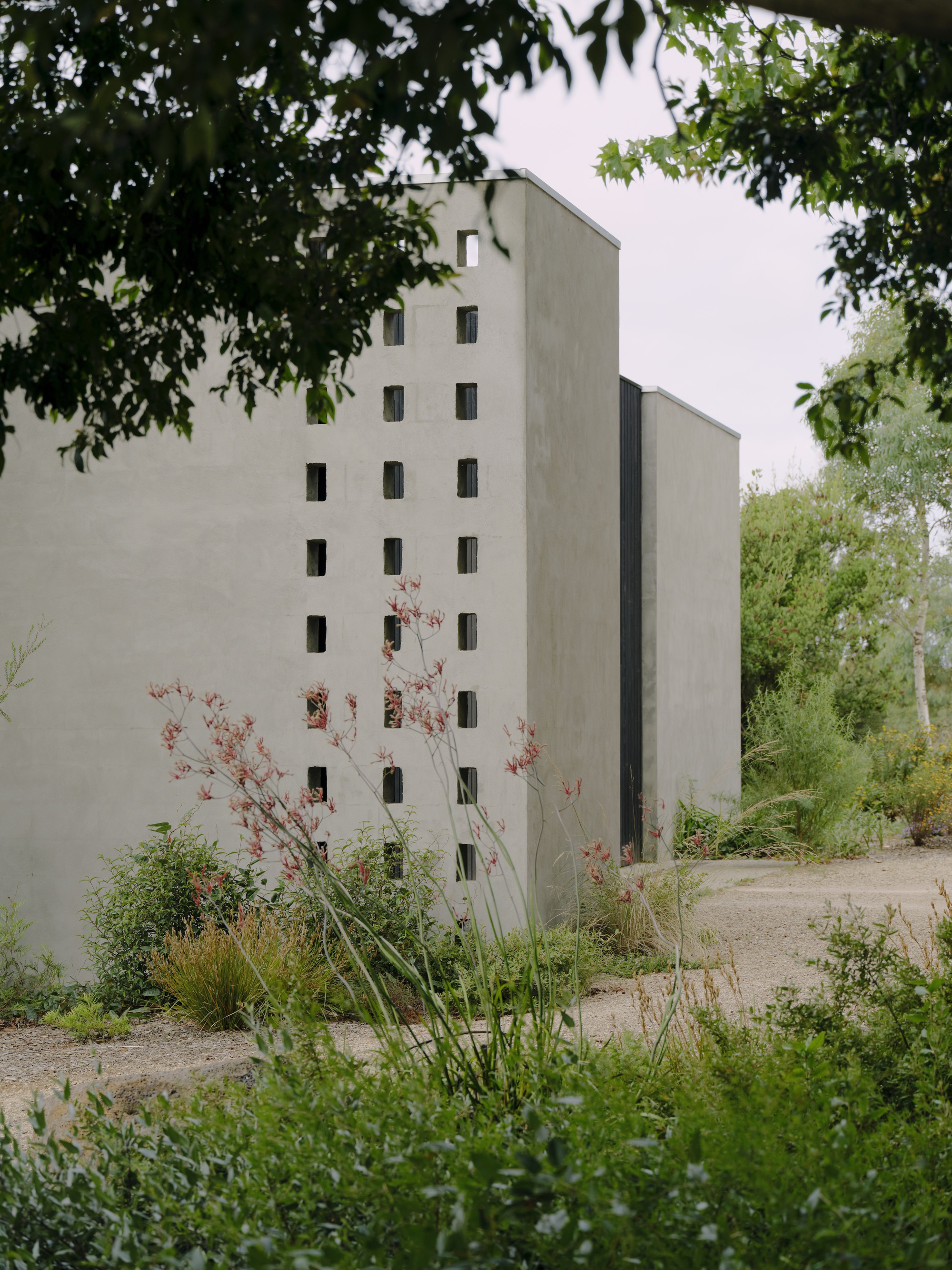 Tall concrete tower with vertical window pattern rising above garden beds with grasses and shrubs