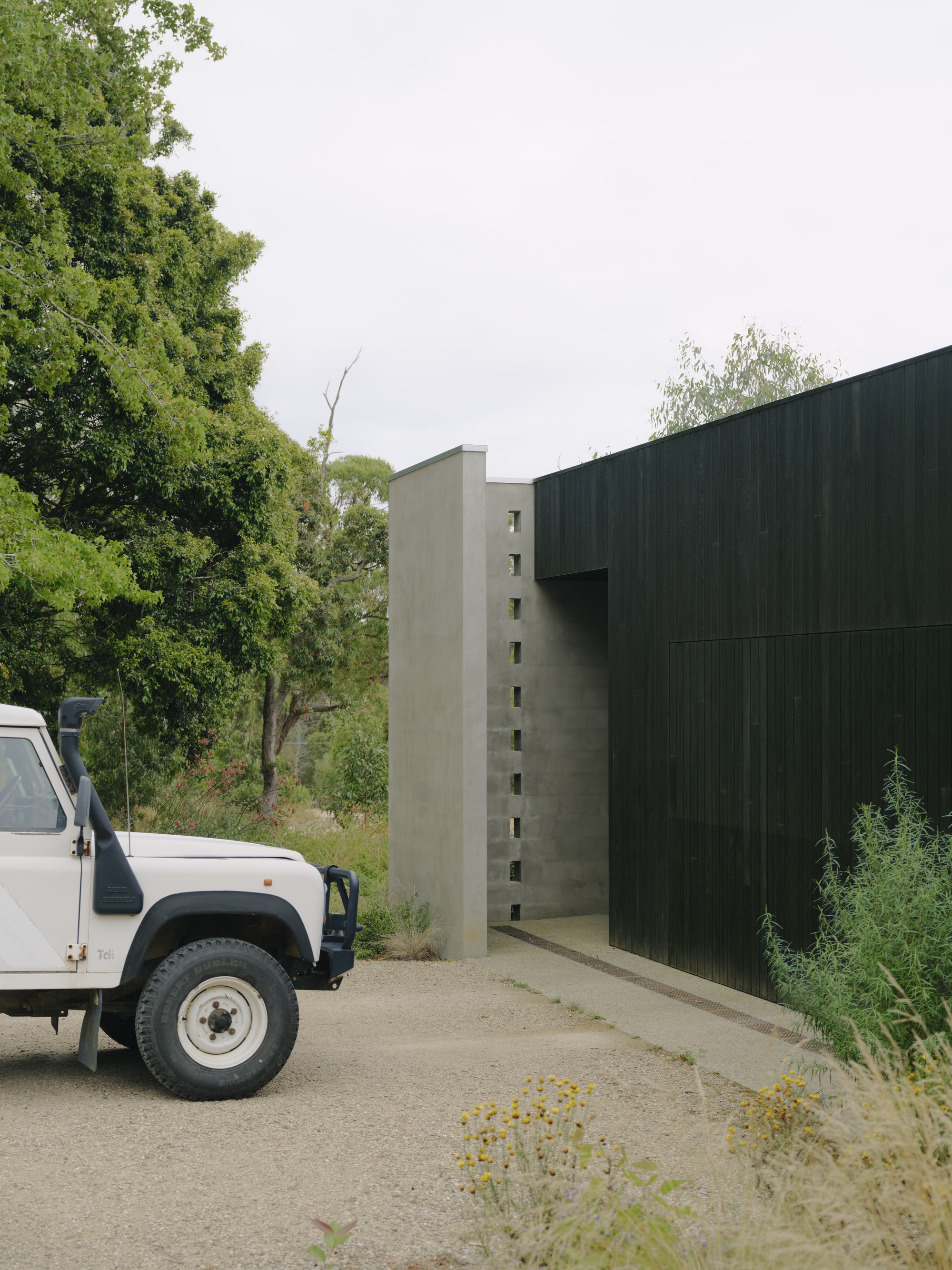 Concrete wall with vertical window openings meeting corrugated metal cladding beside a parked vehicle