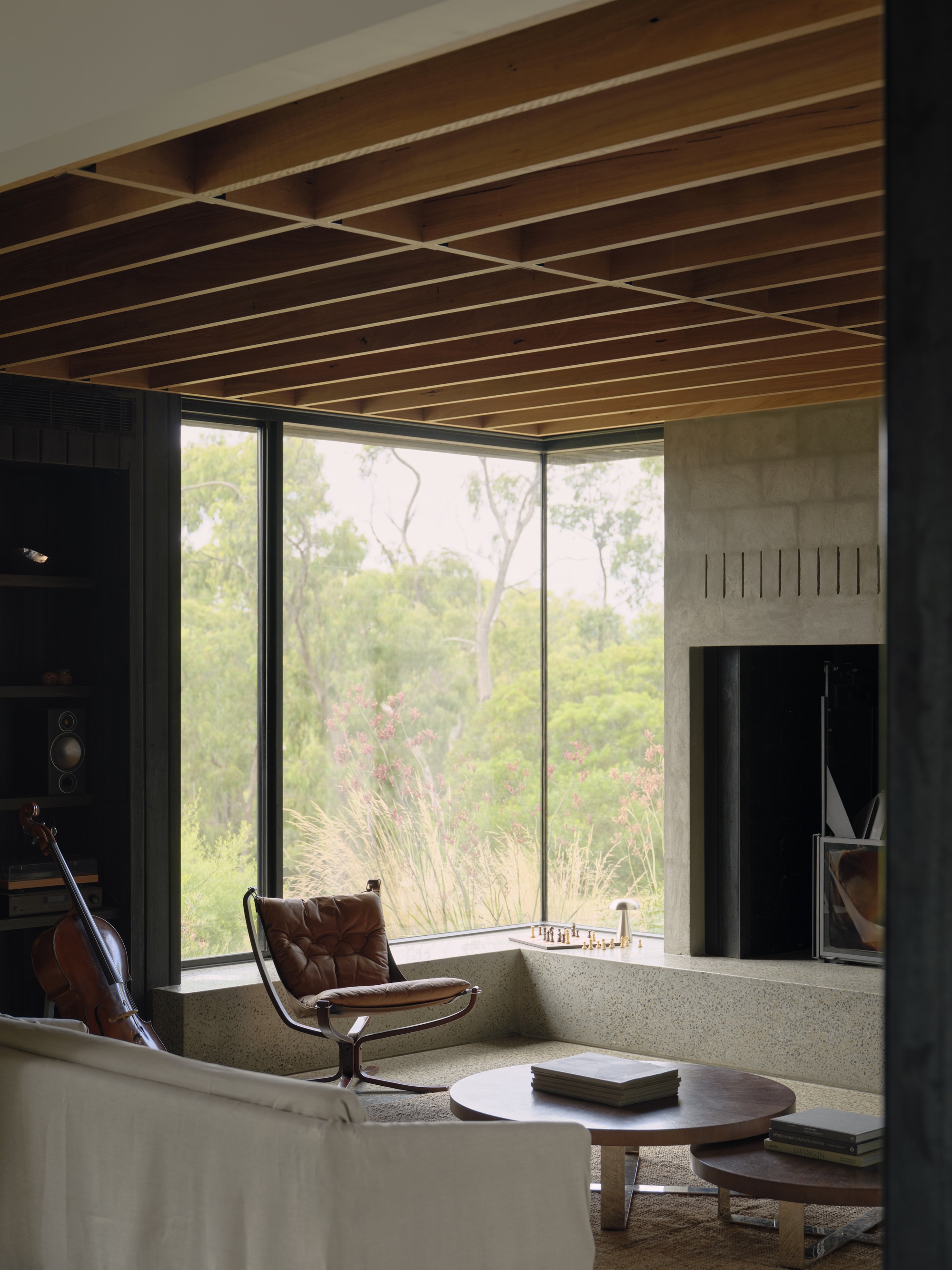 Living room with timber slat ceiling and floor-to-ceiling windows overlooking bushland with cello beside fireplace