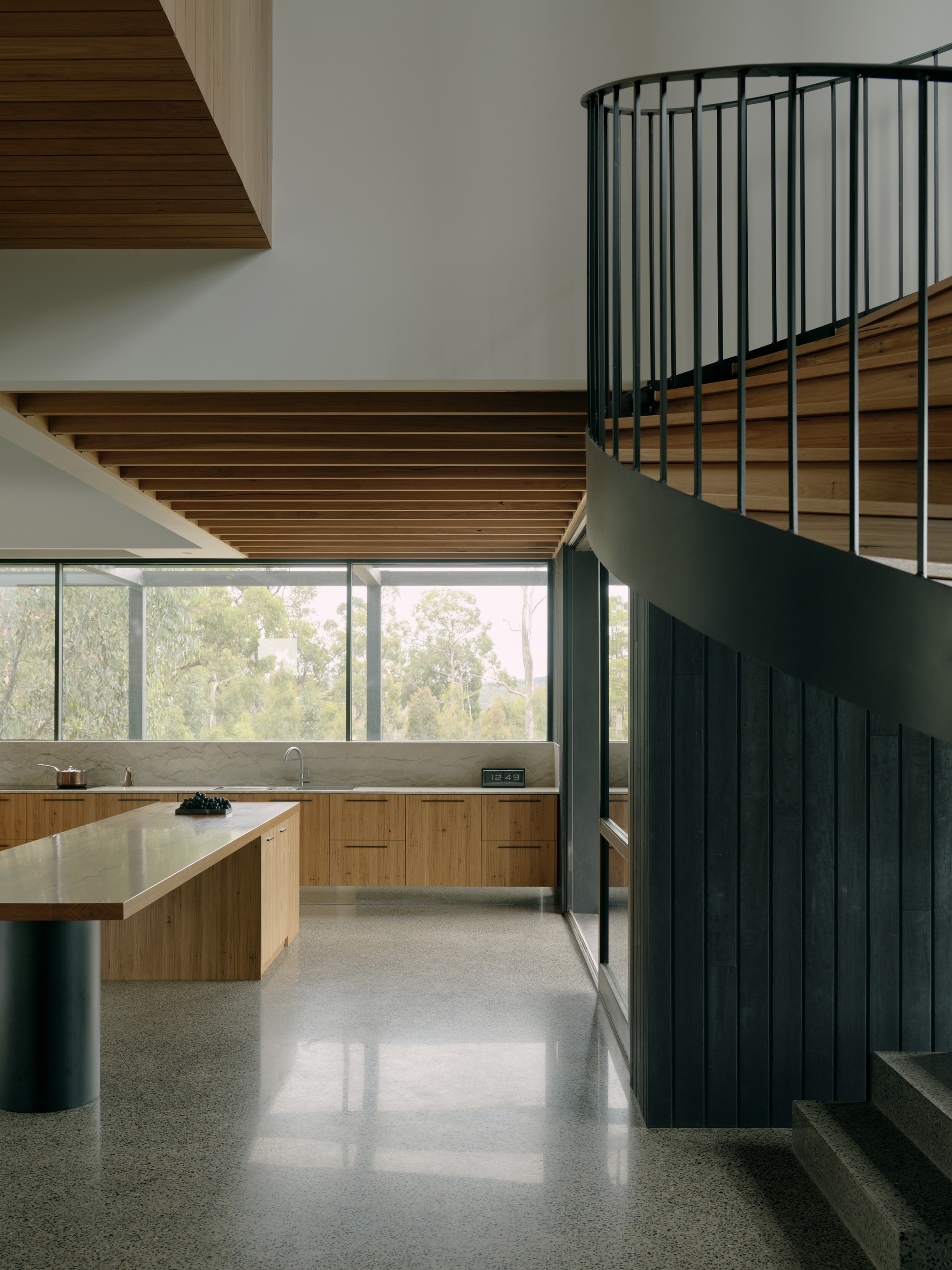 Curved steel staircase with vertical balustrade rising through kitchen with timber slat ceiling and terrazzo flooring