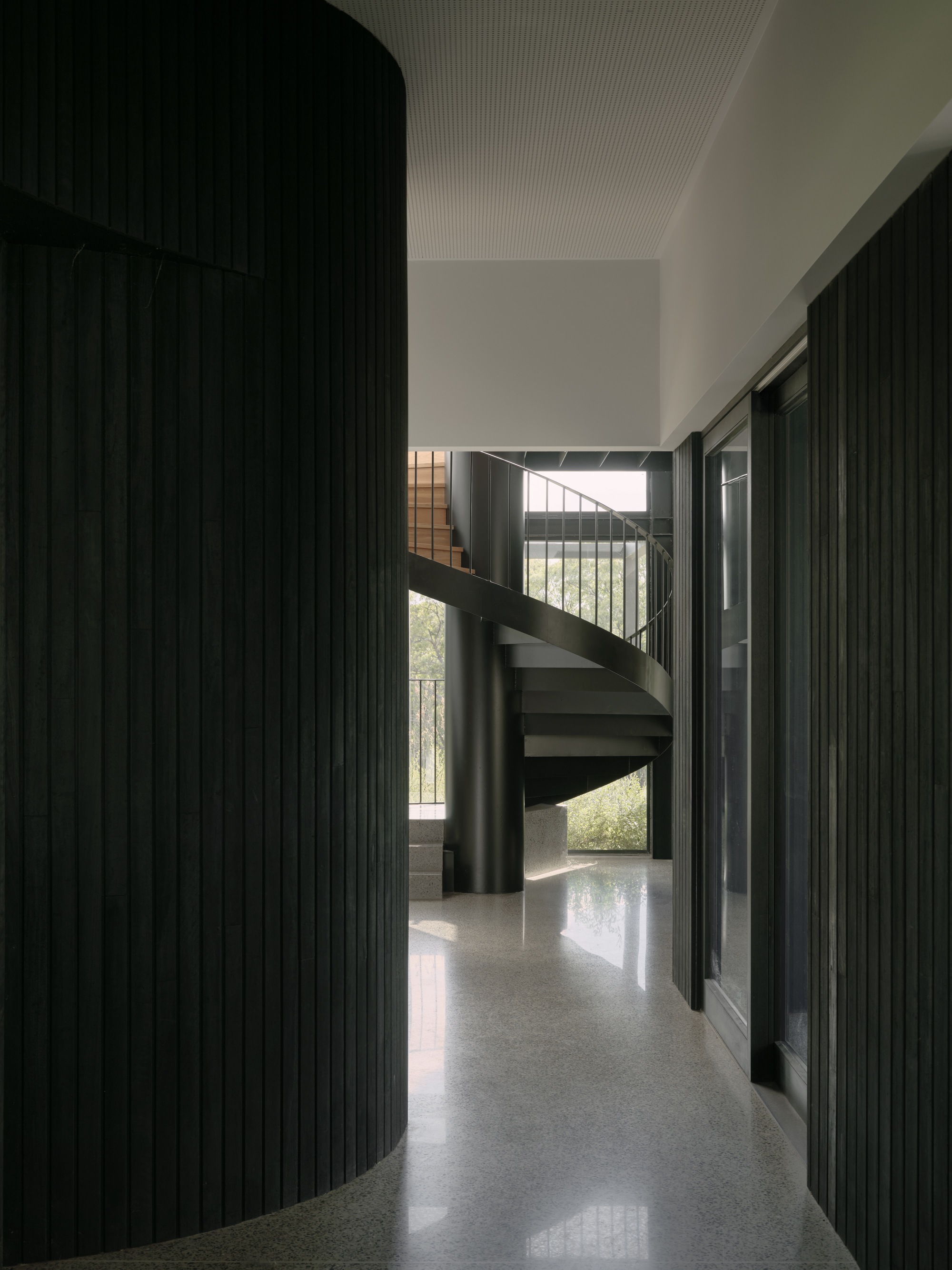 Curved black timber-clad wall beside a spiral staircase in a corridor with polished concrete floors