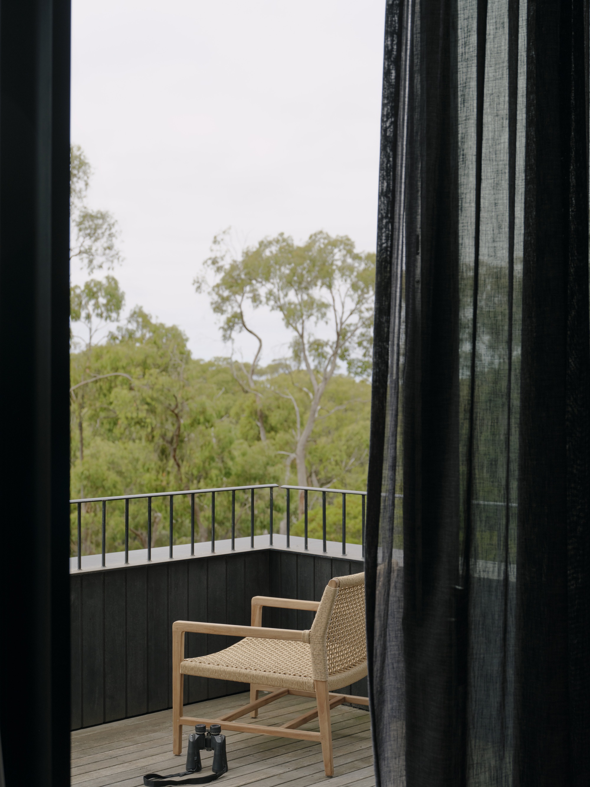 Balcony with woven timber chair and binoculars glimpsed through sheer black curtains