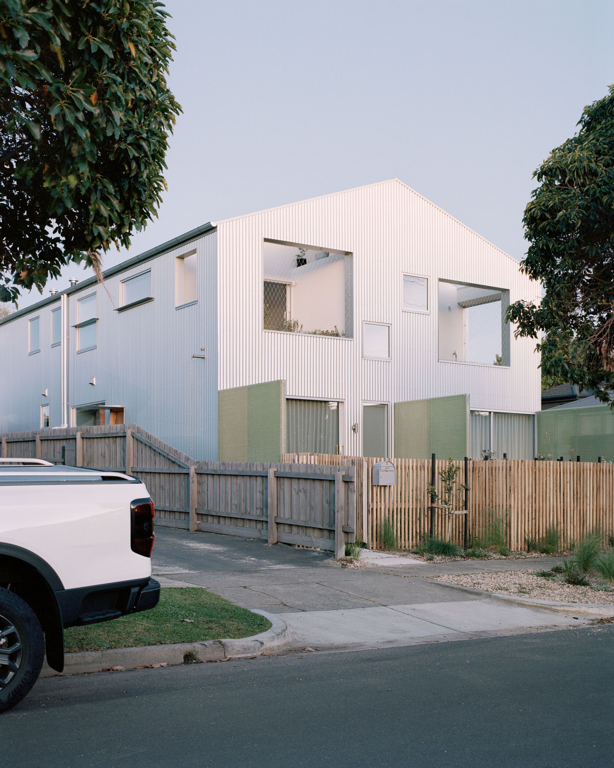 Street view of the white corrugated metal facade with timber fencing and foliage at dusk