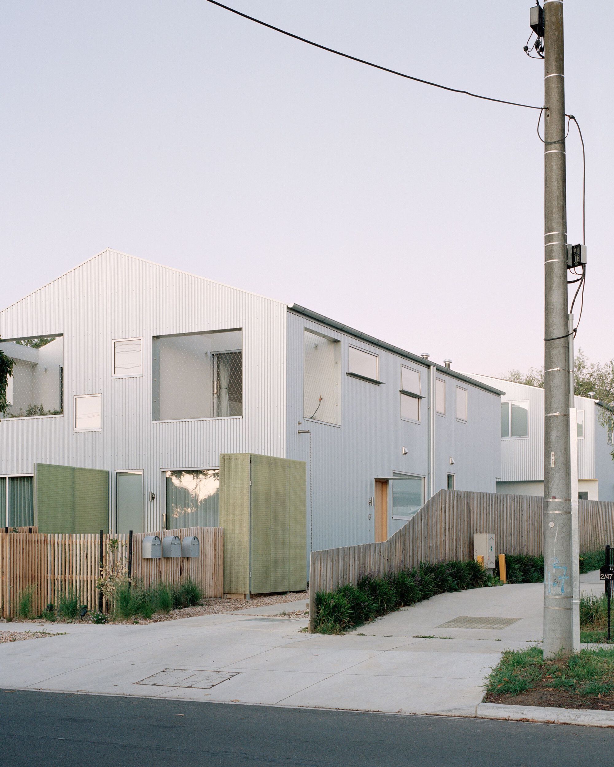 Street elevation showing the gabled corrugated volumes with timber fencing and a utility pole