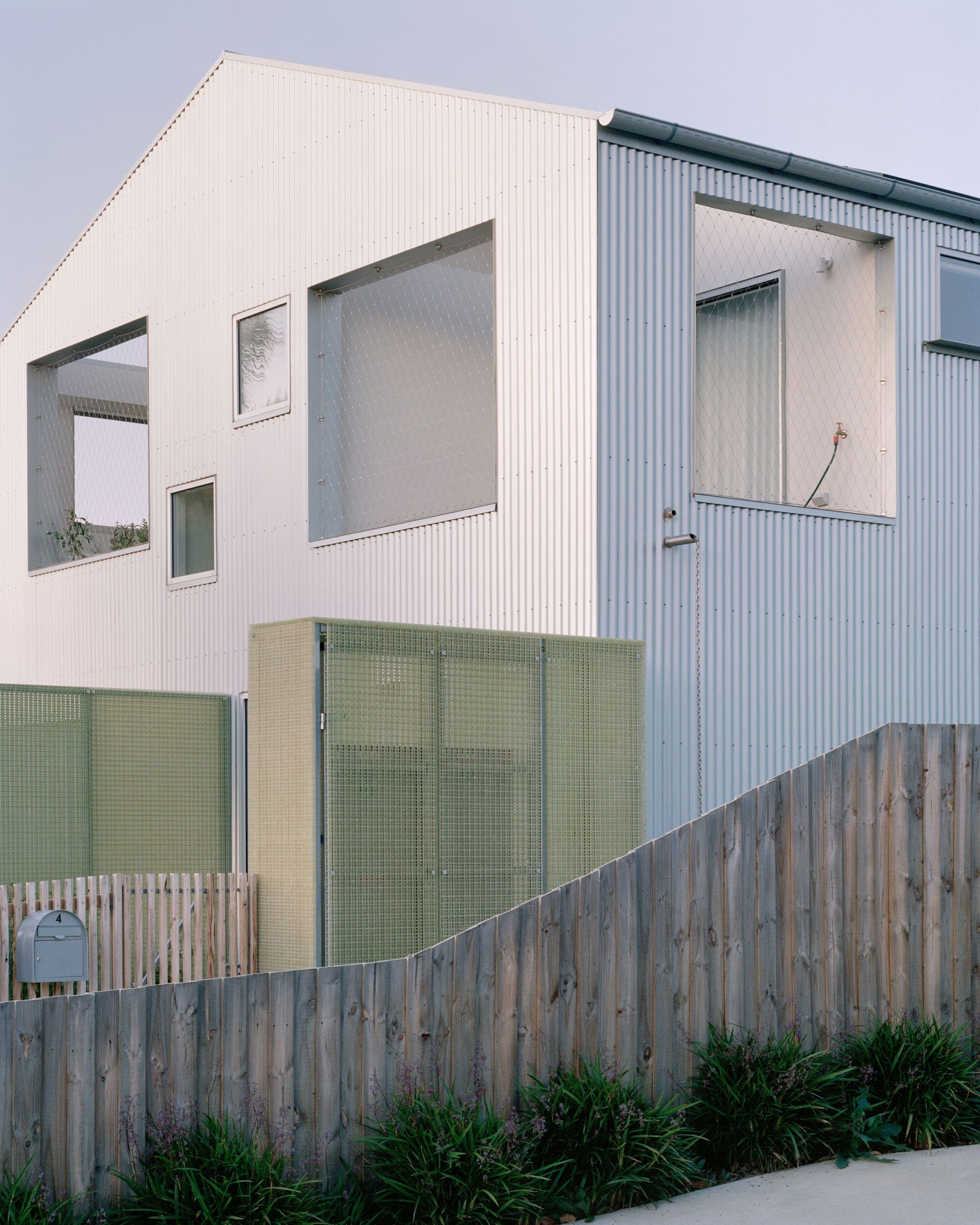 Street view of white corrugated metal facade with green mesh screening behind a timber fence
