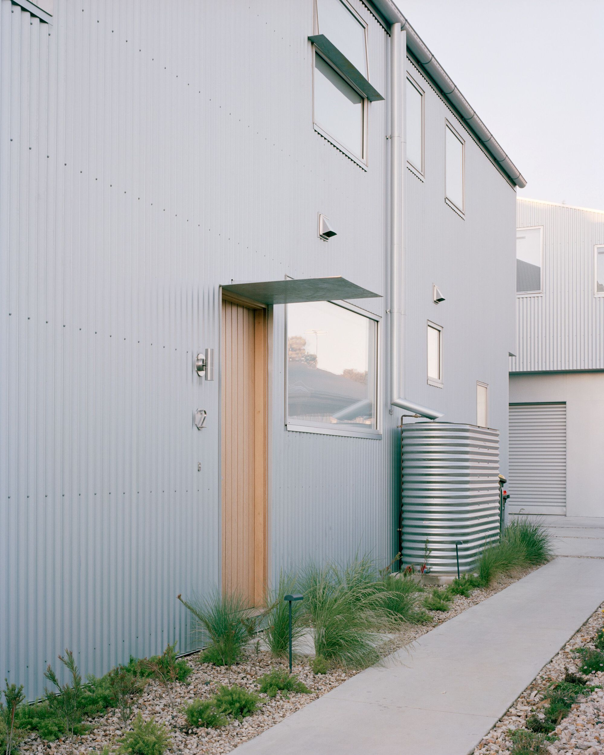 Entry facade with timber door surround and corrugated rainwater tank beside gravel planting bed