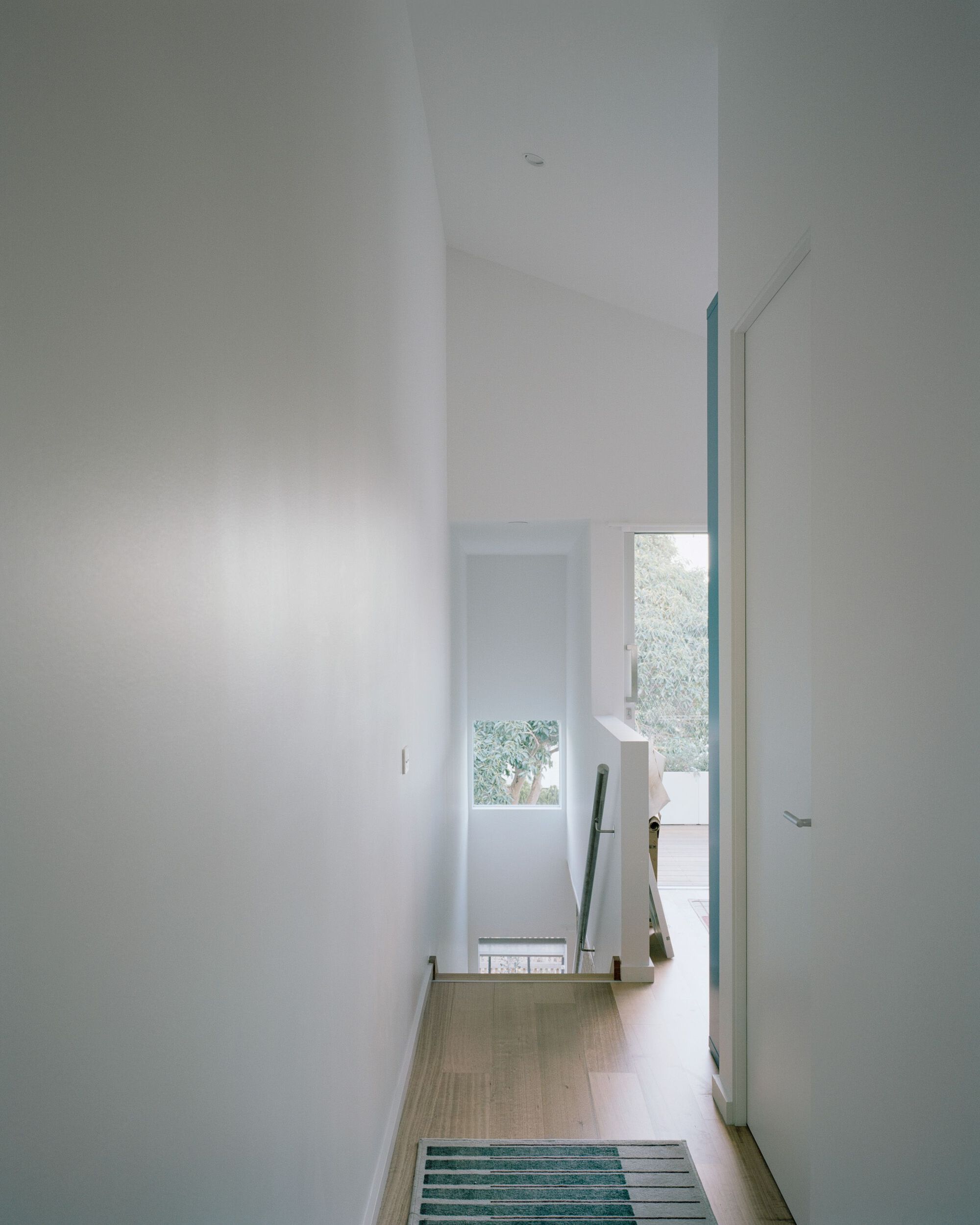 Narrow corridor with light wood flooring and a striped blue rug leading to a window with garden view