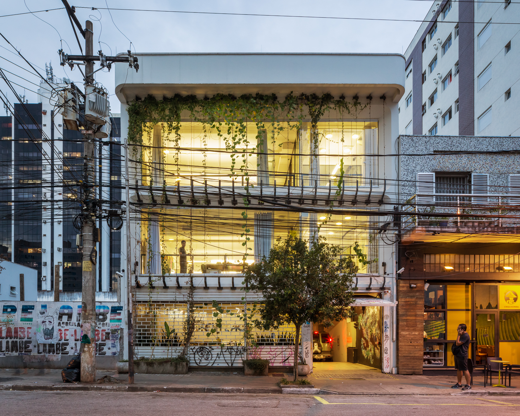 Street facade of a two-storey glass-fronted building draped in climbing vines, lit warmly at dusk behind a tangle of overhead wires