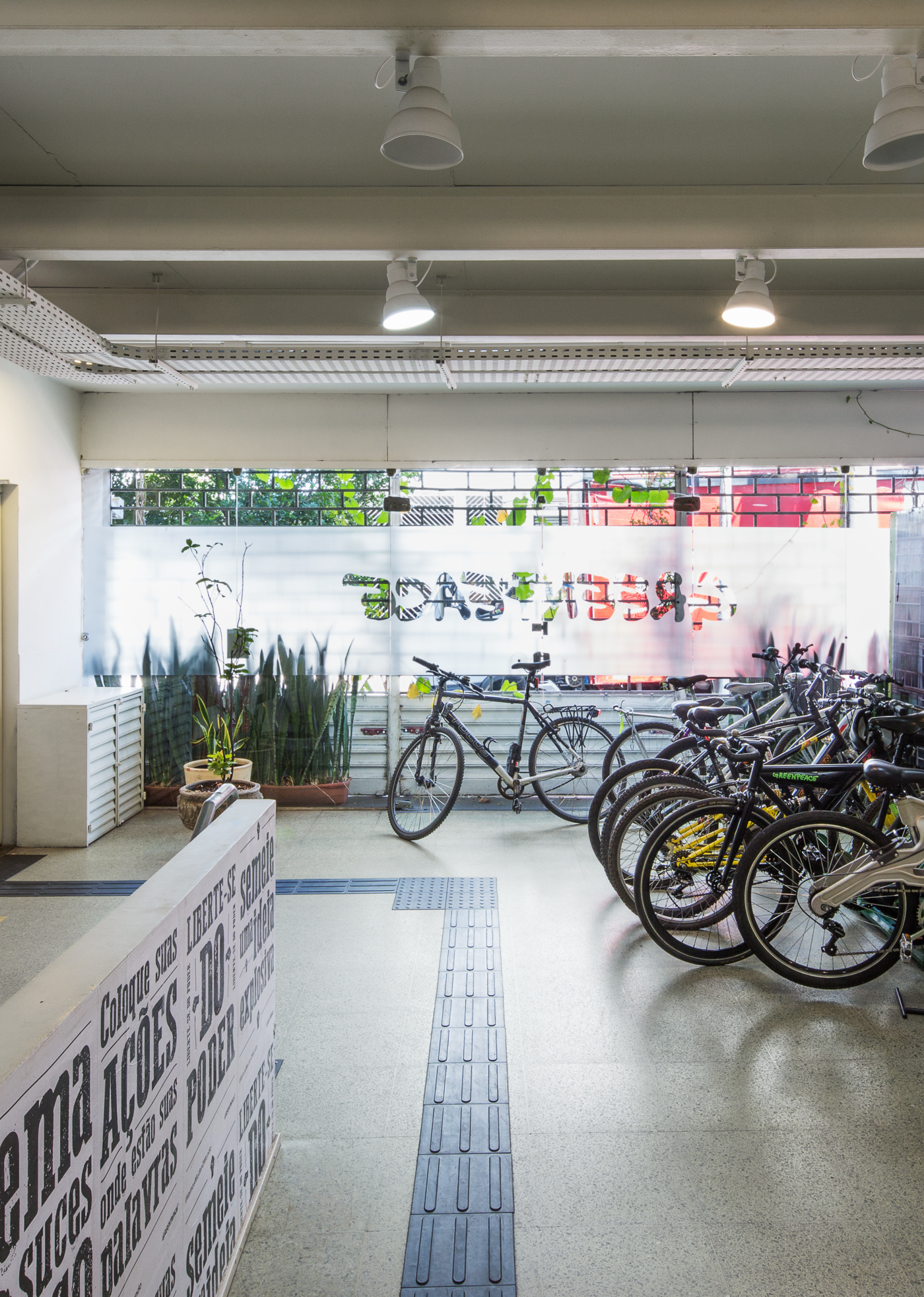 Ground-floor bicycle parking with a tactile paving strip, potted plants and a typographic wall under spotlights in daylight