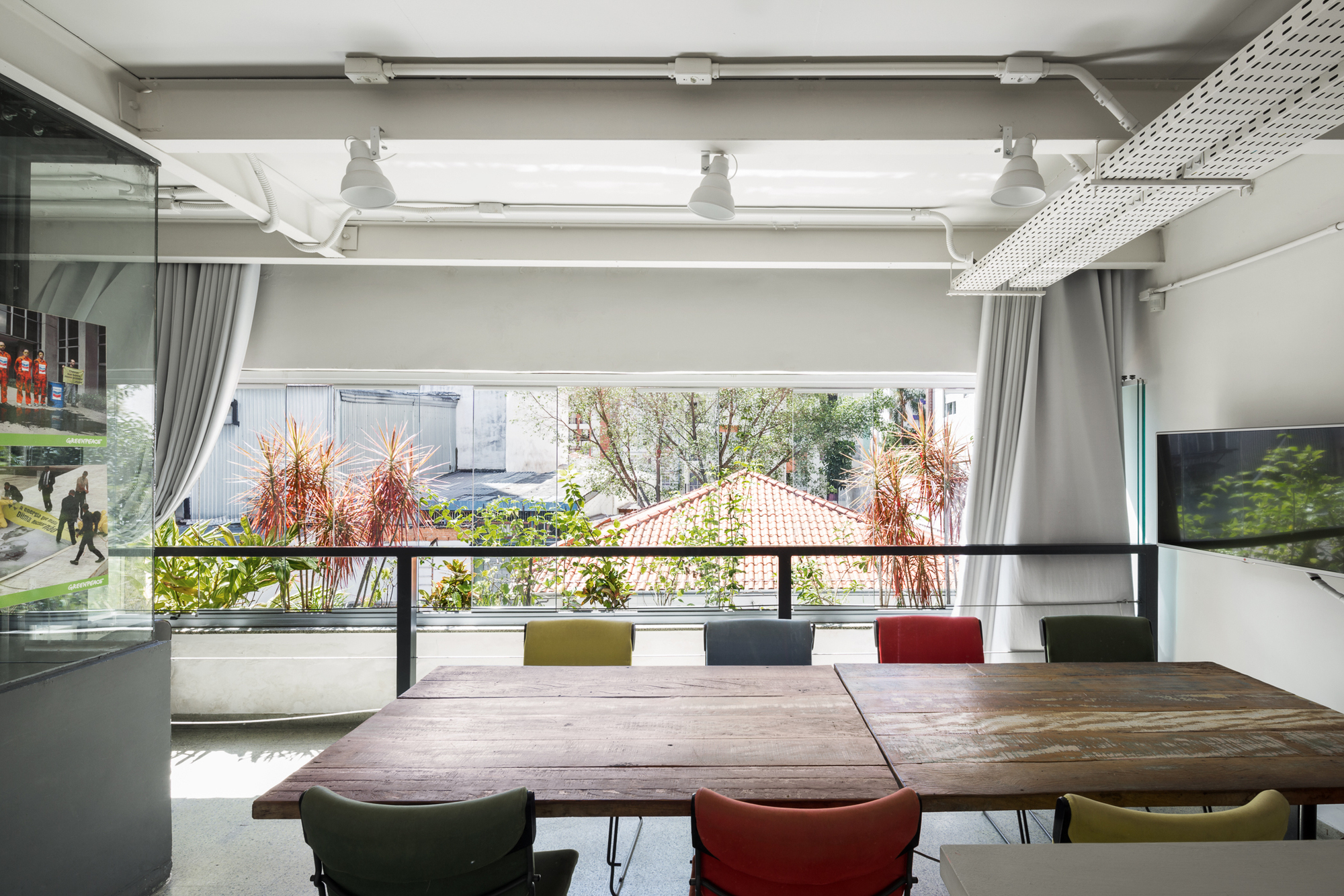 Meeting table with mismatched colourful chairs facing a balcony of red-leaved plants and a tiled roof beyond, under exposed ceiling services