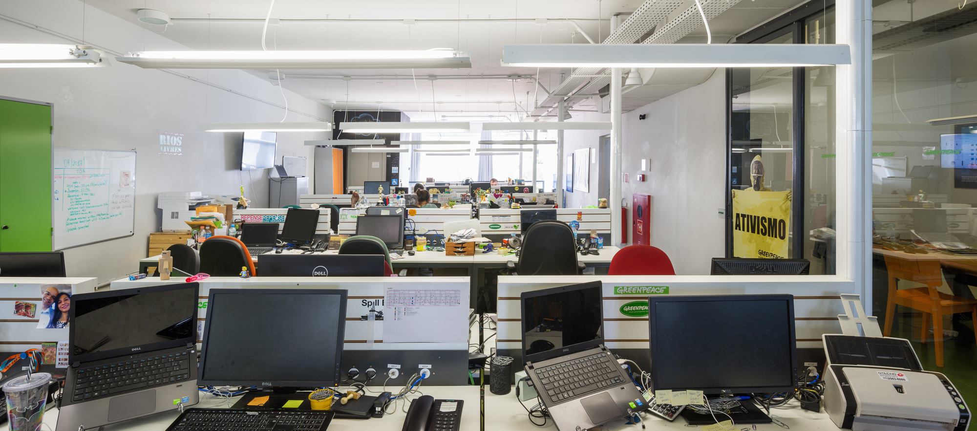 Open-plan office with rows of desks, monitors and laptops under linear pendant lights, with a glazed meeting room at right