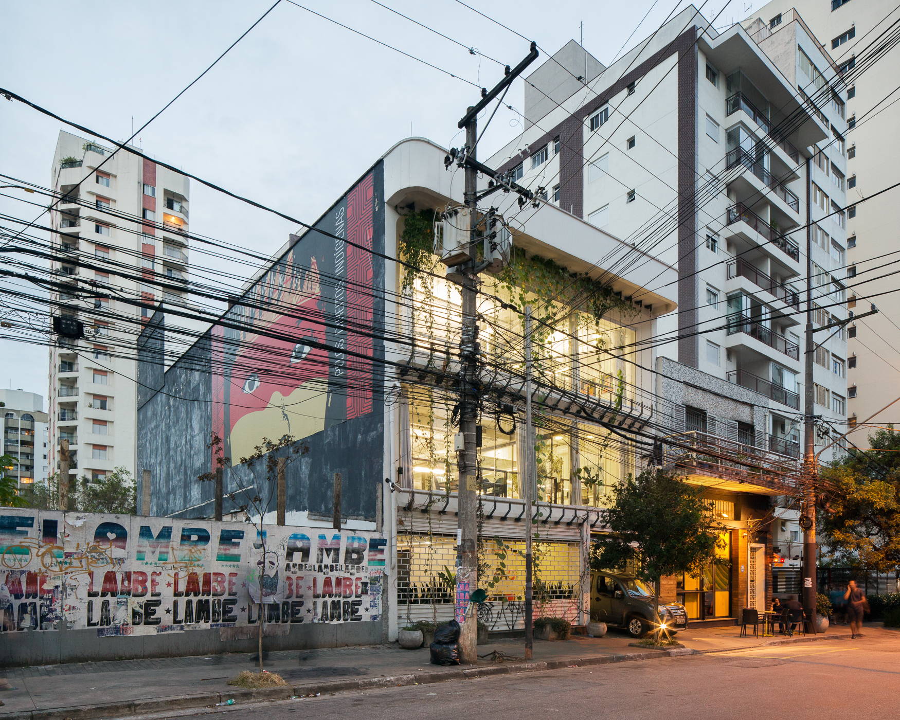 Street corner view of the glazed, vine-draped building beside a large mural, with utility wires crossing the dusk sky