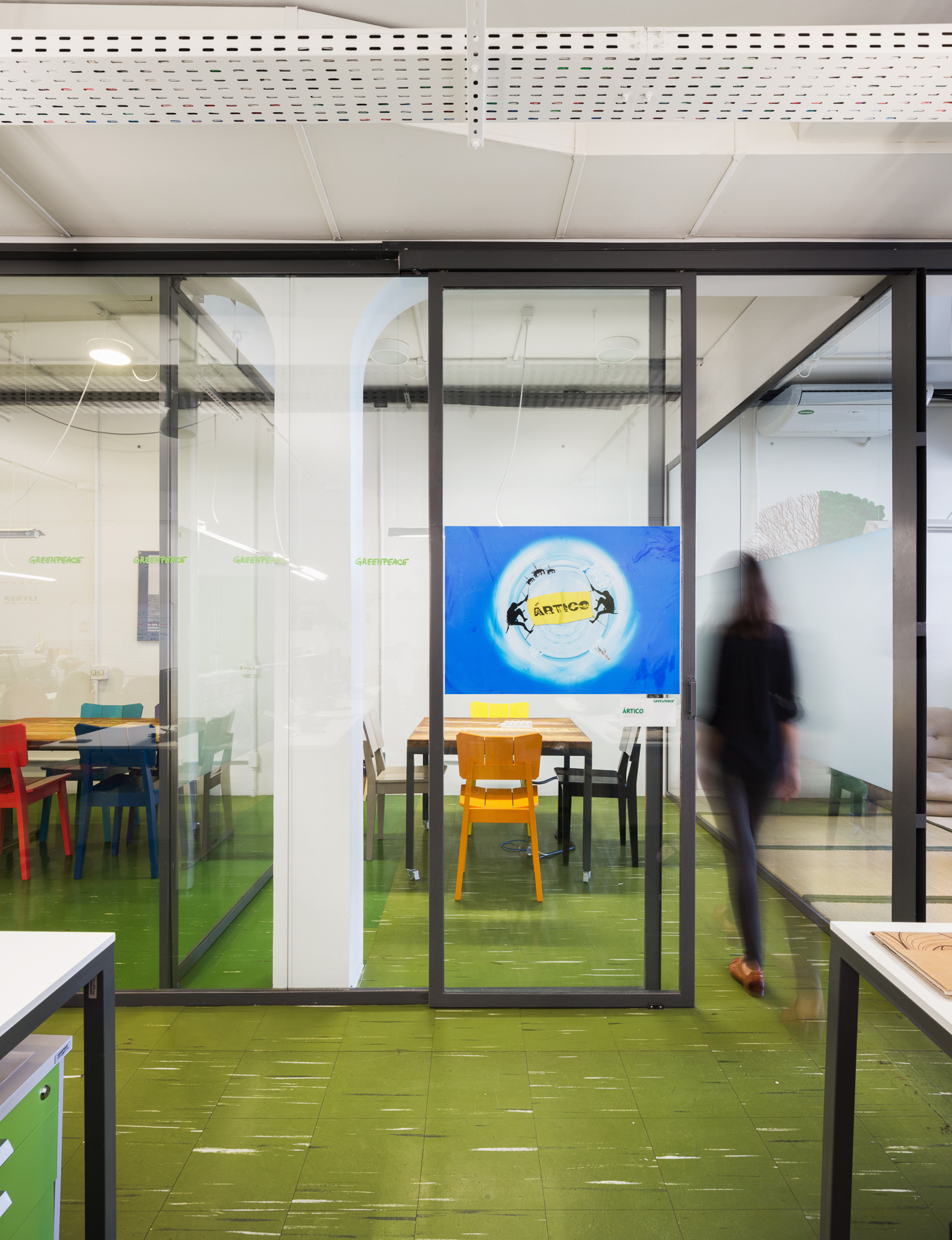 Glazed meeting room with orange and red chairs seen across a green floor, a blurred person walking past