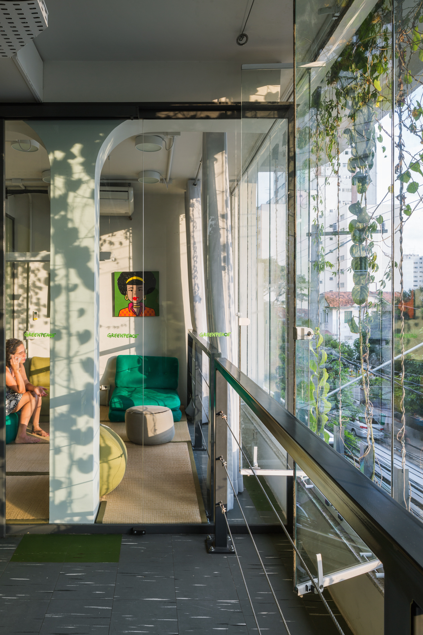 Glass-walled meeting nook with a green armchair and framed portrait, seen past a cable railing in late afternoon sun