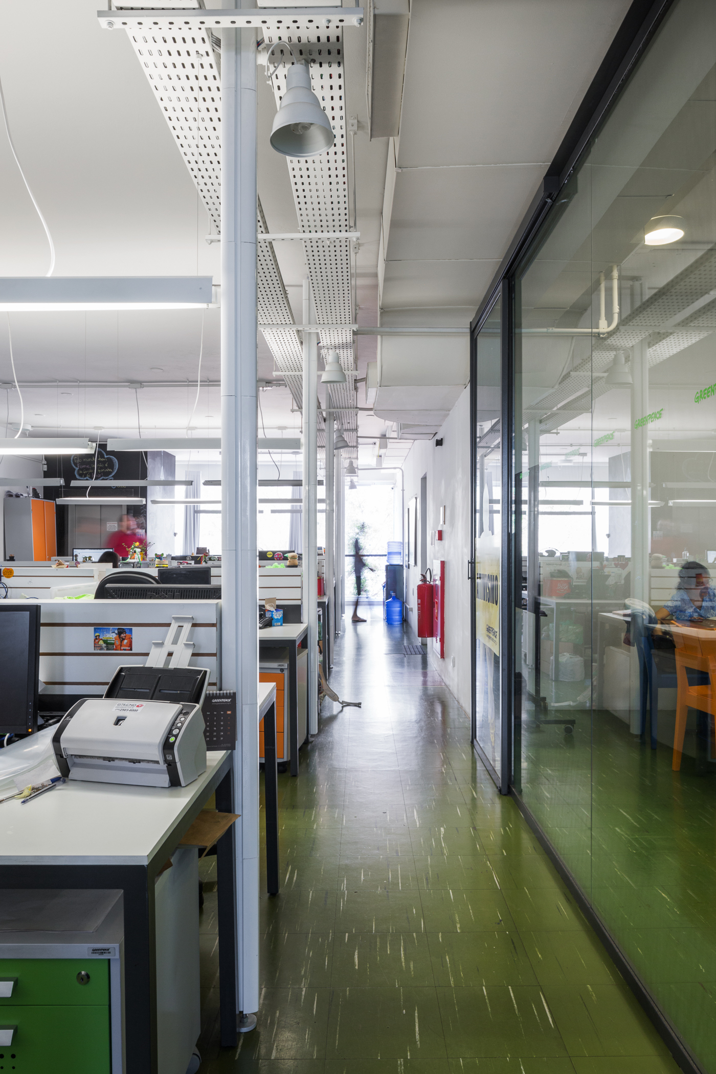 Long open-plan office corridor with green speckled floor, white columns and a glazed partition reflecting a seated worker