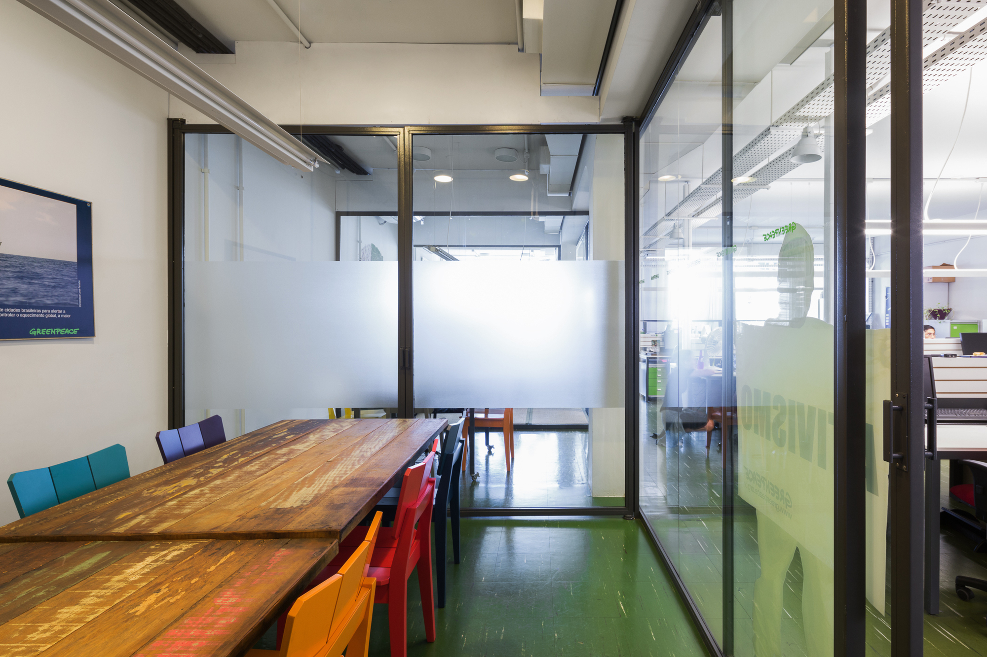 Meeting room with a reclaimed timber table, colorful chairs and frosted glass partitions over a green floor