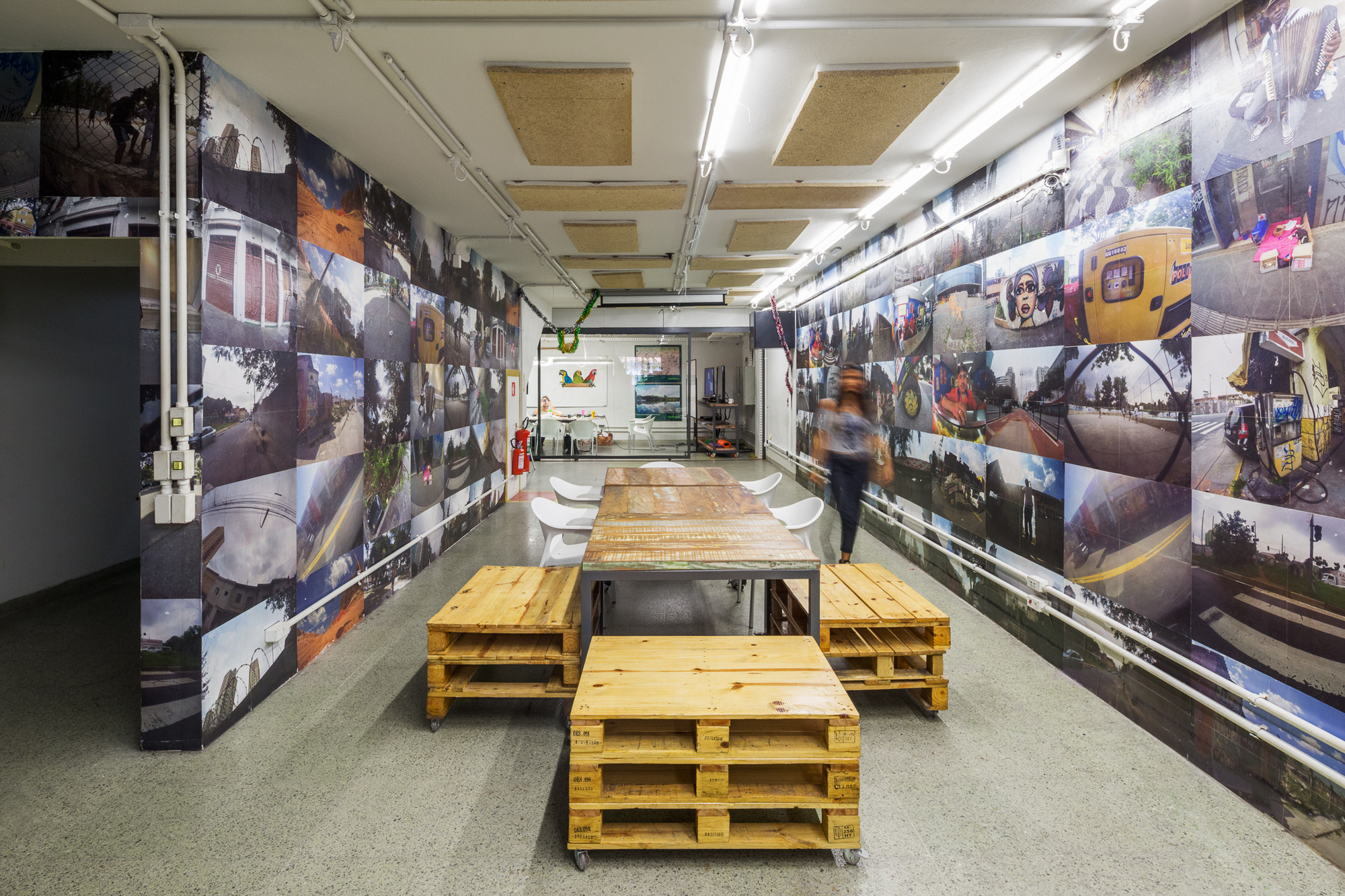 Corridor-like meeting space lined with large photo murals, pallet seating and a woman walking past a long table
