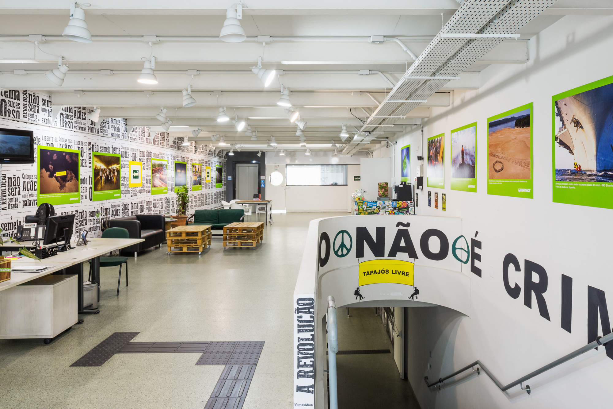 Open office hall with text-covered wall, green-framed photographs, pallet tables and a stairwell with painted lettering