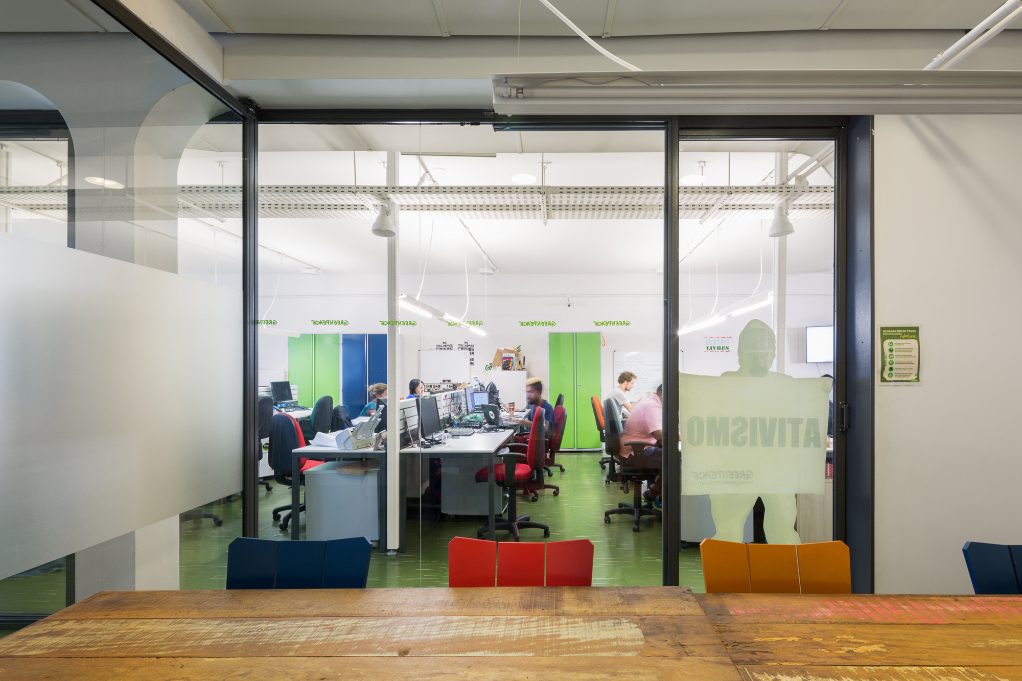 Open-plan office seen through a frosted glass partition, with workers at shared desks and red chairs on a green floor