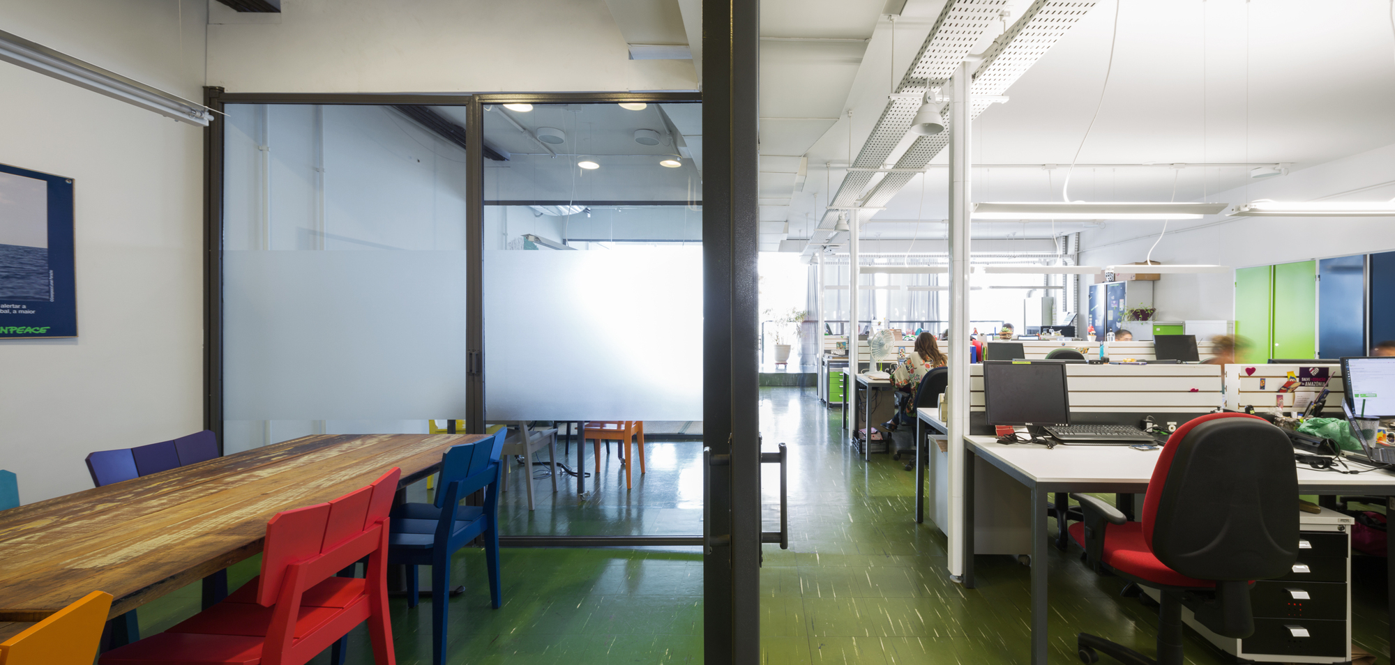 Long view along the green-floored office past a reclaimed timber table with colored chairs and a glass partition