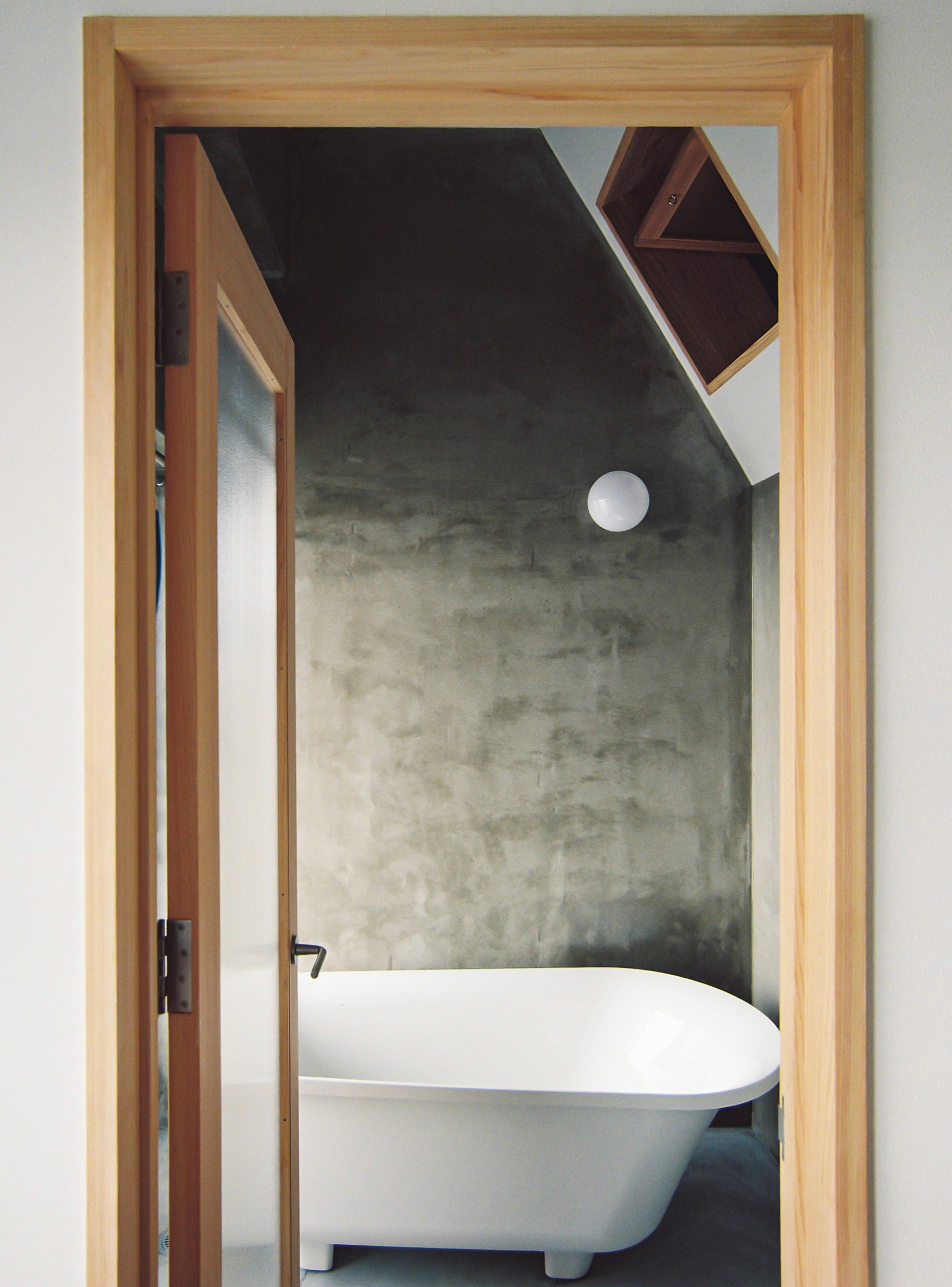 Freestanding bathtub framed by timber doorway beneath tilted concrete ceiling plane with circular skylight