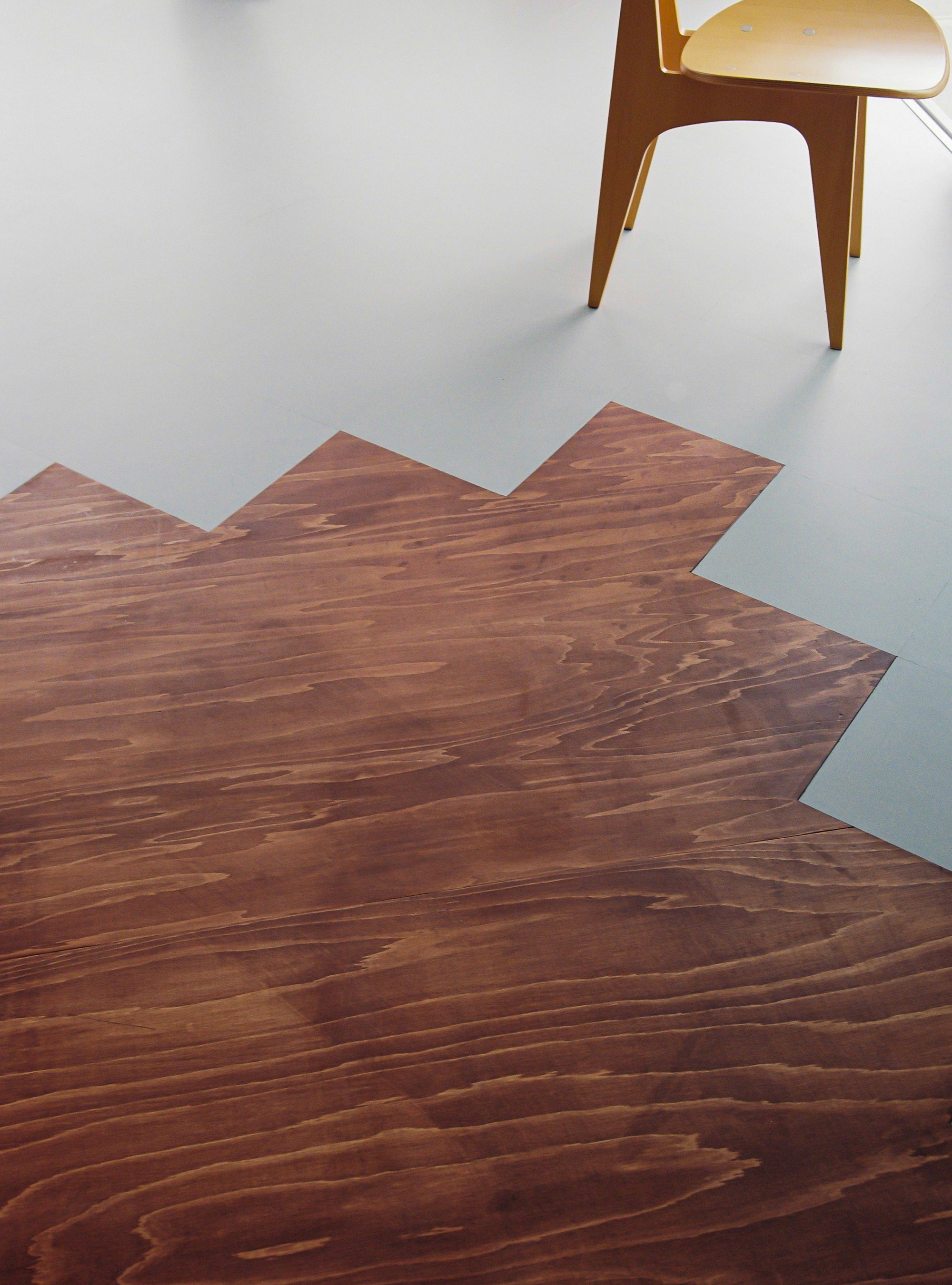 Overhead view of zigzag wood floor edge meeting pale linoleum with timber chair legs visible