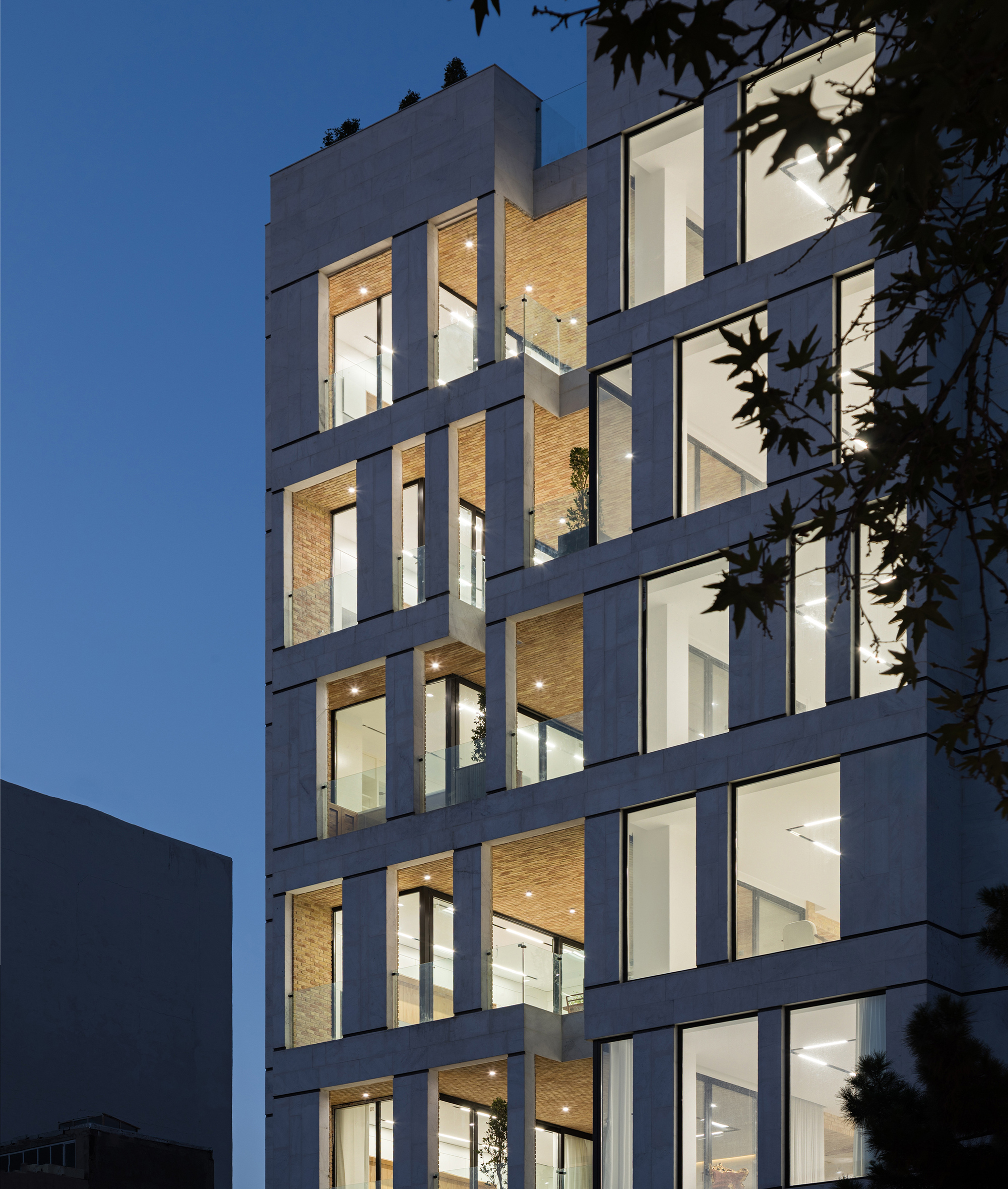 Illuminated facade with timber-lined balconies at dusk framed by tree branches