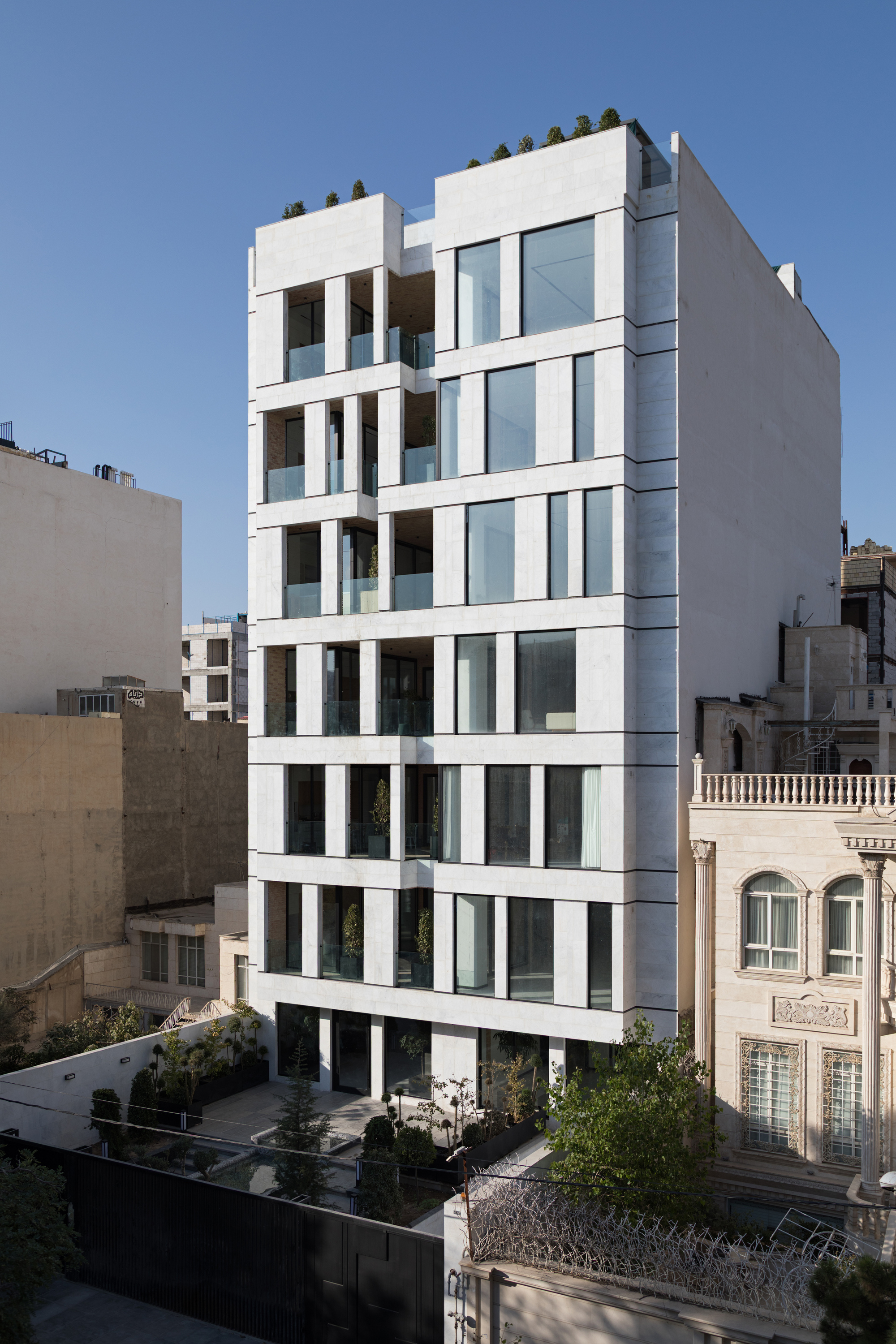 White facade with recessed balconies and floor-to-ceiling glazing among neighboring residential buildings