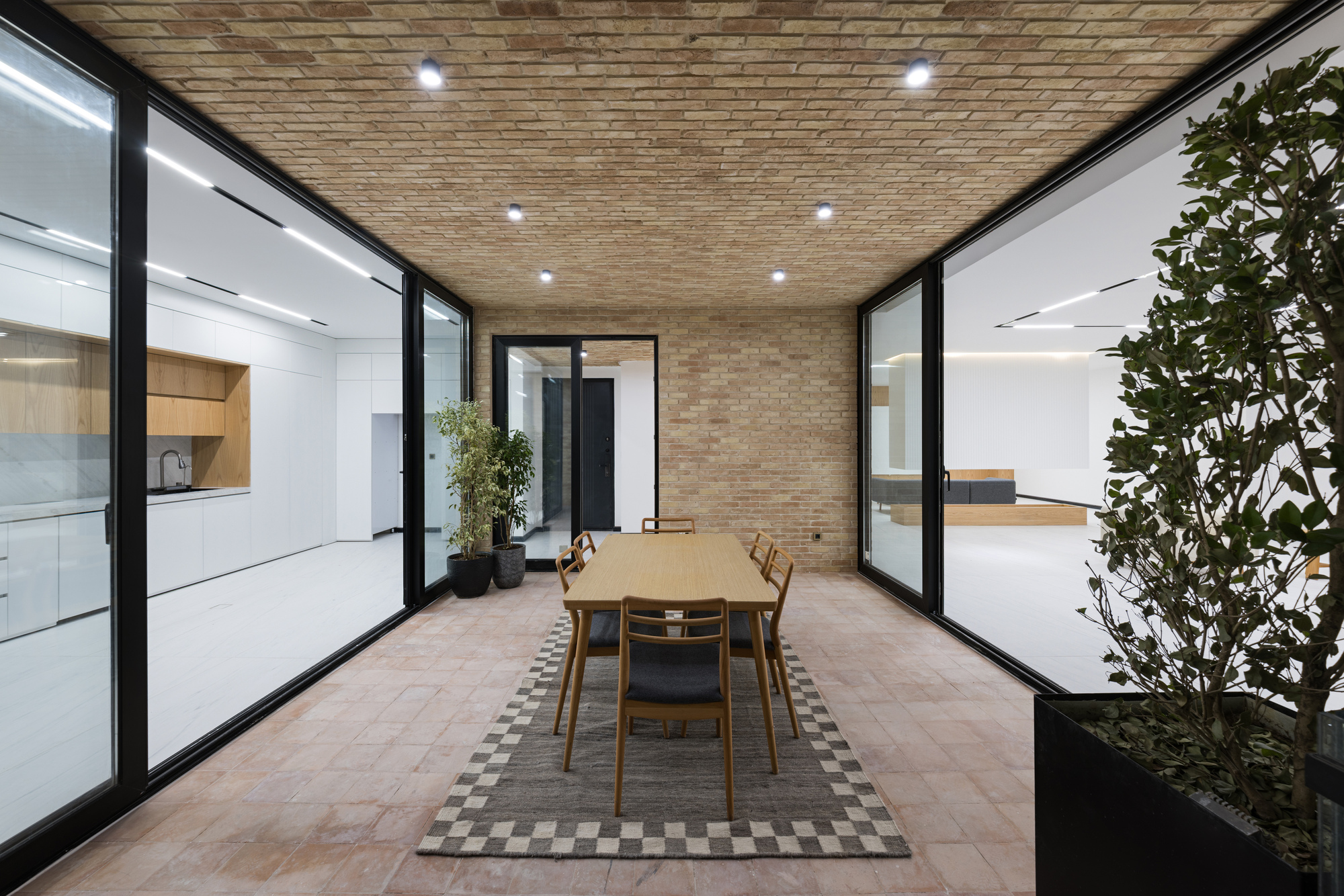 Terrace with dining table flanked by potted trees and illuminated interior spaces at dusk
