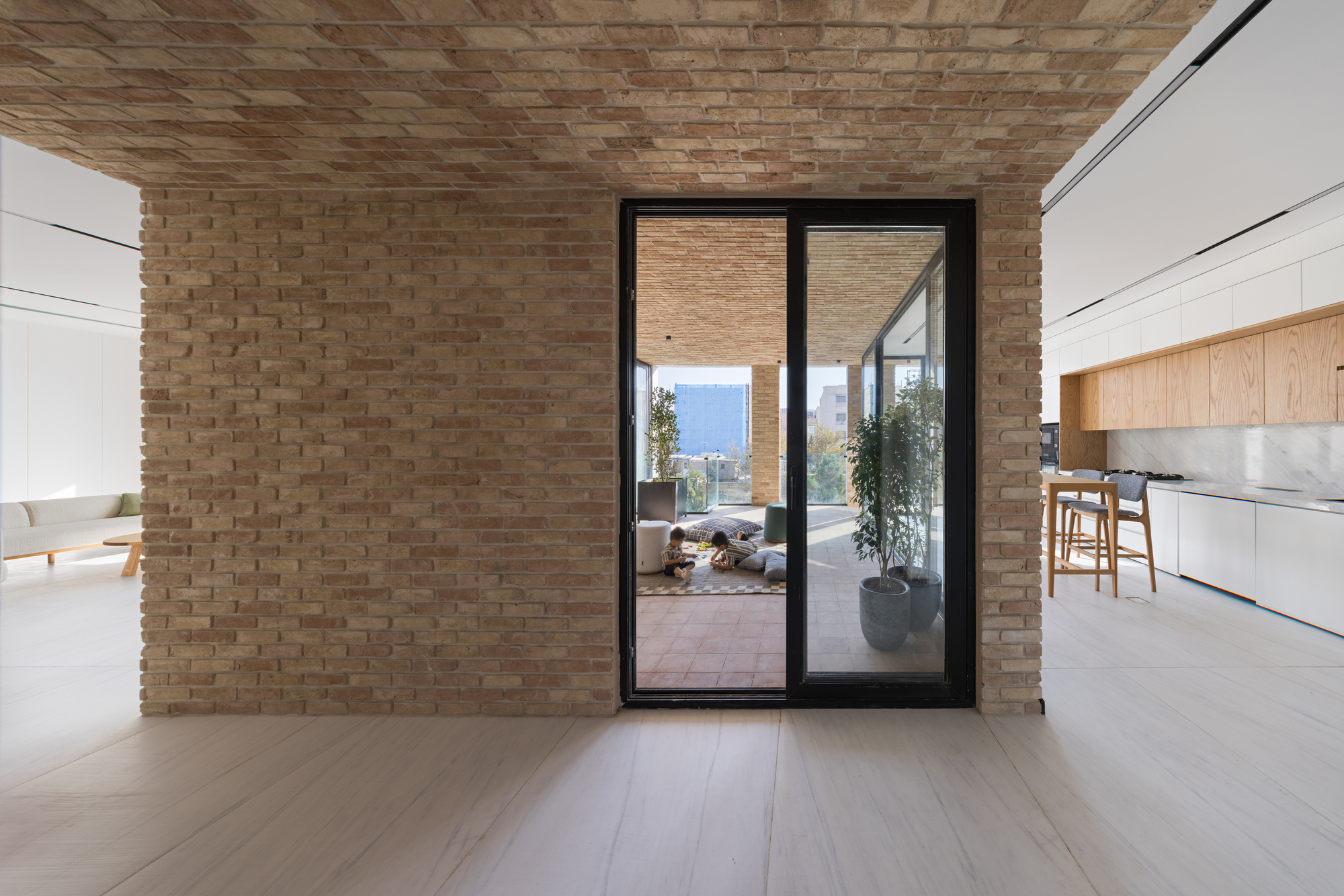 Interior view through black-framed glass doors to an exterior terrace with woven brick ceiling and potted plants