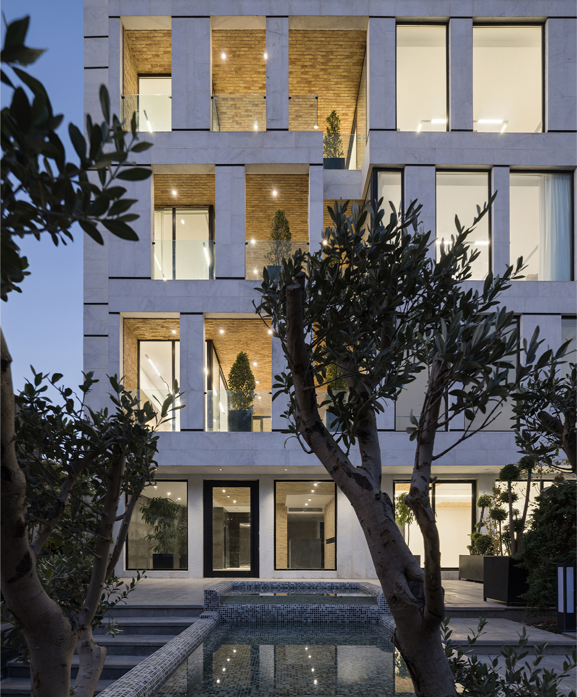 Street facade at dusk showing staggered brick balconies framed by concrete slabs behind mature olive trees
