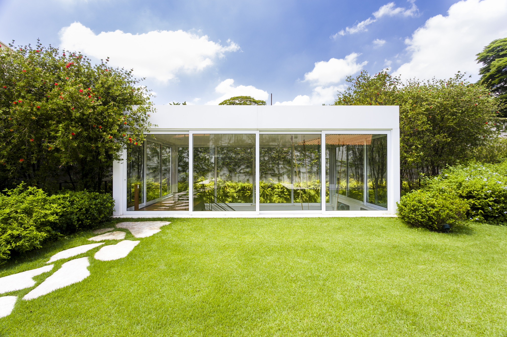 Glazed white box pavilion facing a lawn with stepping stones, flanked by flowering shrubs under a blue sky with clouds