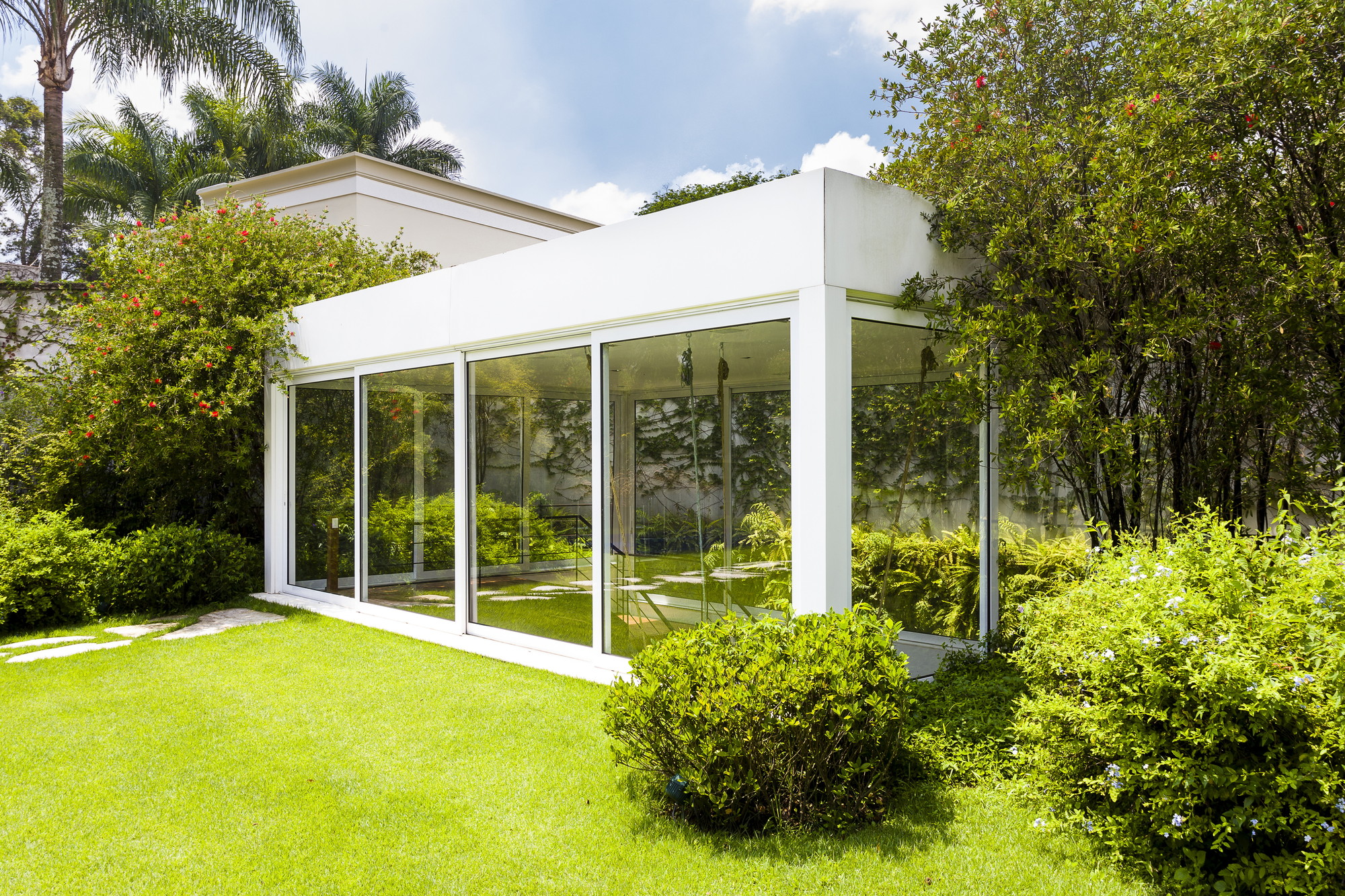 Corner of the glass-walled pavilion with a white flat roof, framed by clipped shrubs and red-flowering trees in midday sun