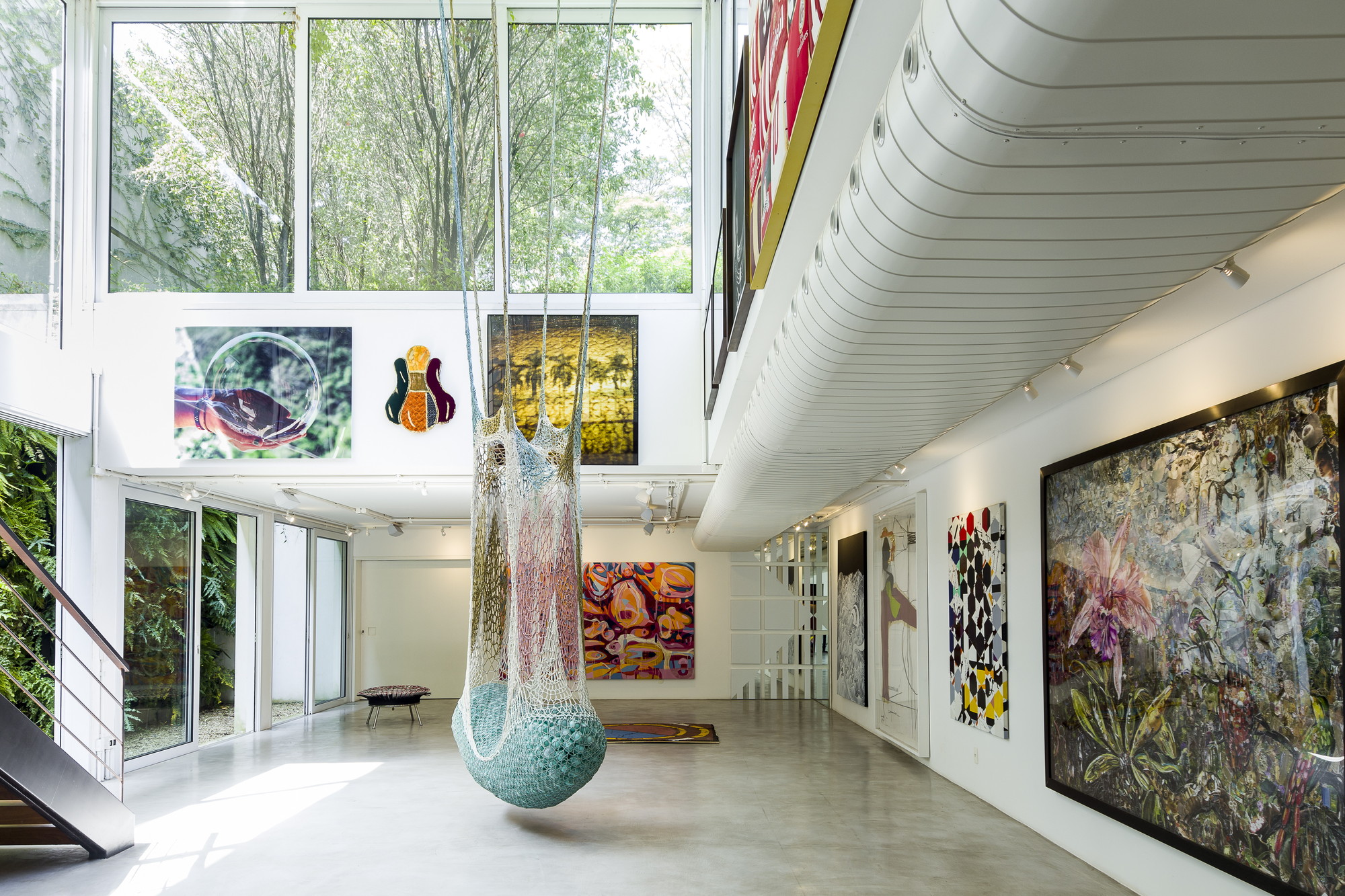 Double-height gallery with a hanging crocheted net sculpture, framed paintings and a curved white soffit, lit by daylight from tall windows