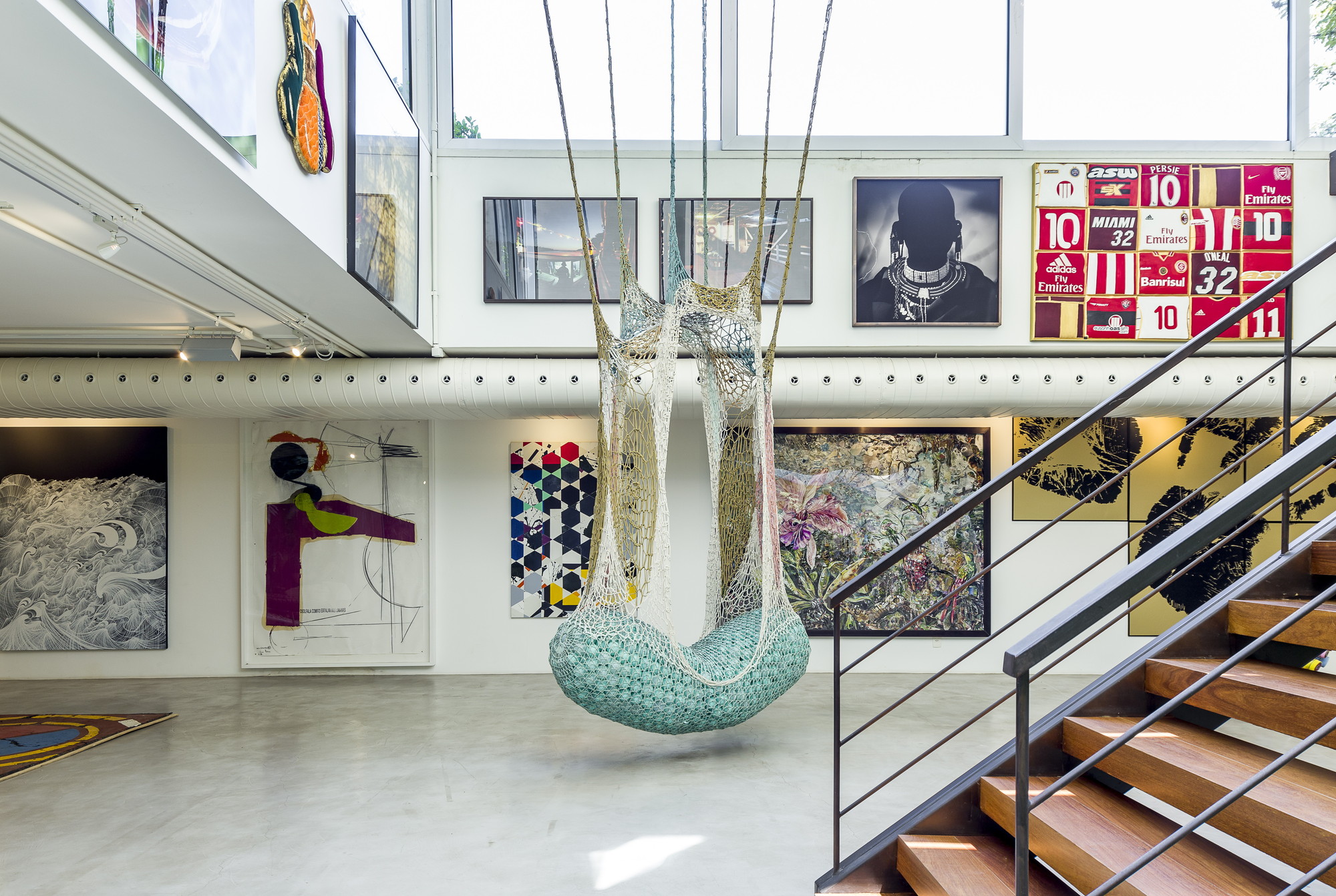 Woven net hammock chair with a teal base hanging in a double-height gallery beside a timber-tread staircase