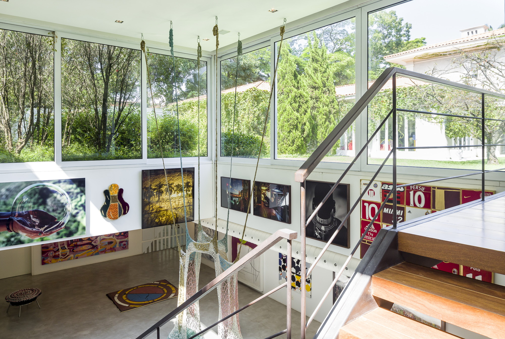 Corner of the double-height gallery with hanging ropes, framed artworks and tall windows looking onto garden trees and a neighboring house