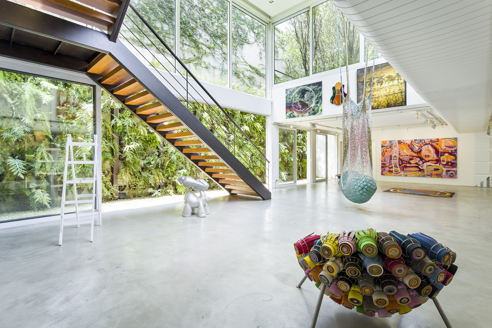 Open gallery floor with a steel and timber staircase rising beside a planted fern wall, a rolled-mat stool in the foreground
