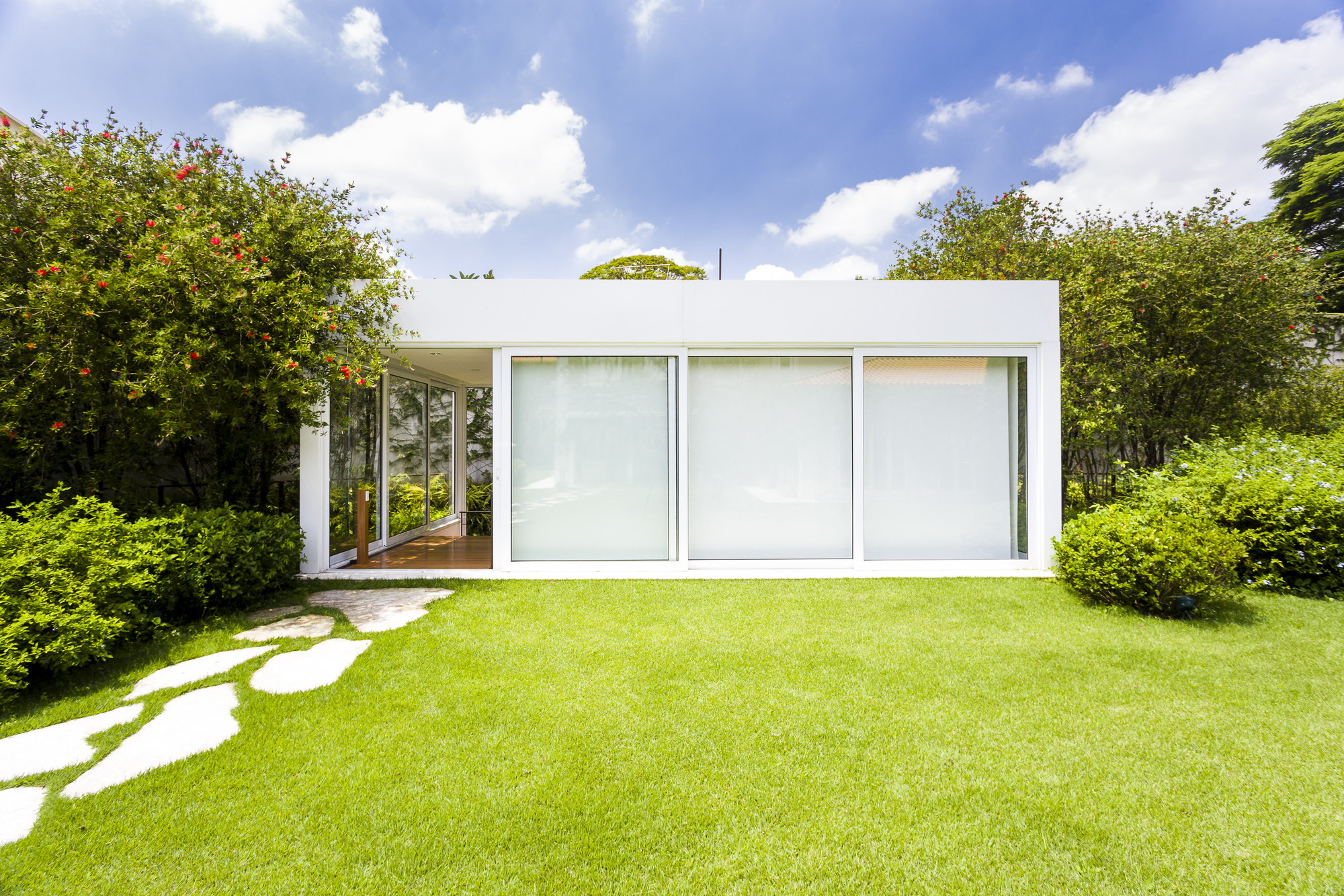 White single-storey pavilion with sliding frosted glass panels, set on a lawn with a stepping-stone path under blue sky