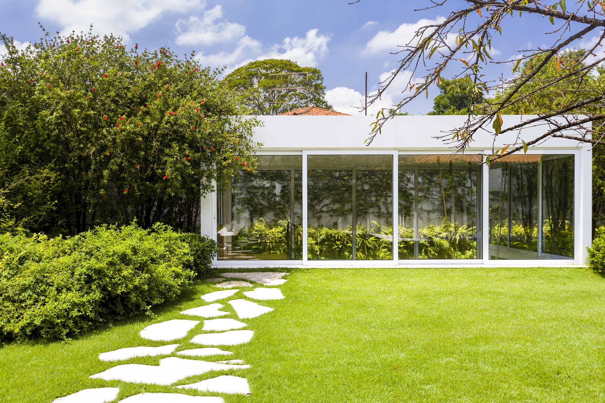 Glazed pavilion reflecting ferns and a vine-covered wall, reached by a stepping-stone path across the lawn on a sunny day