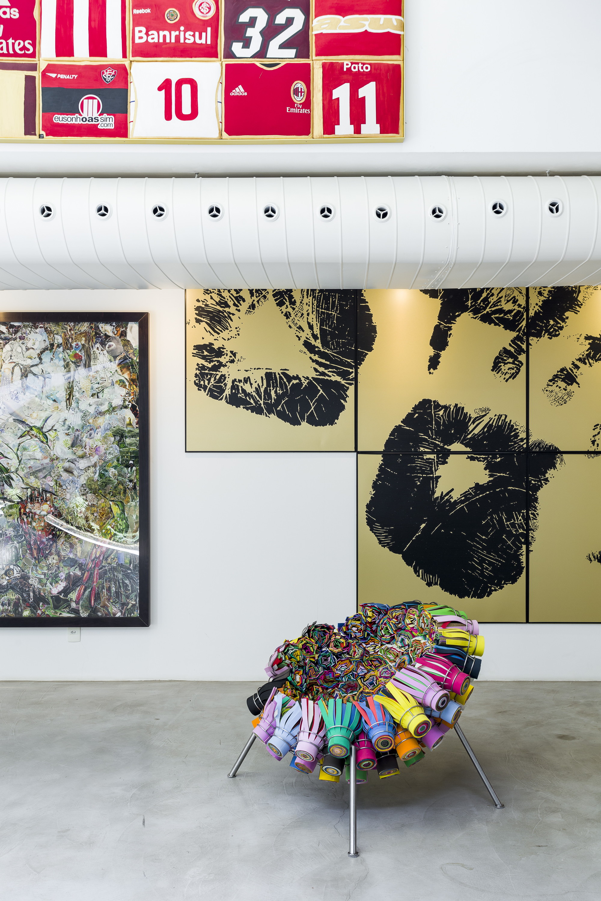 Chair of rolled colourful tubes on steel legs beneath a white duct, framed jerseys and gold lip-print panels
