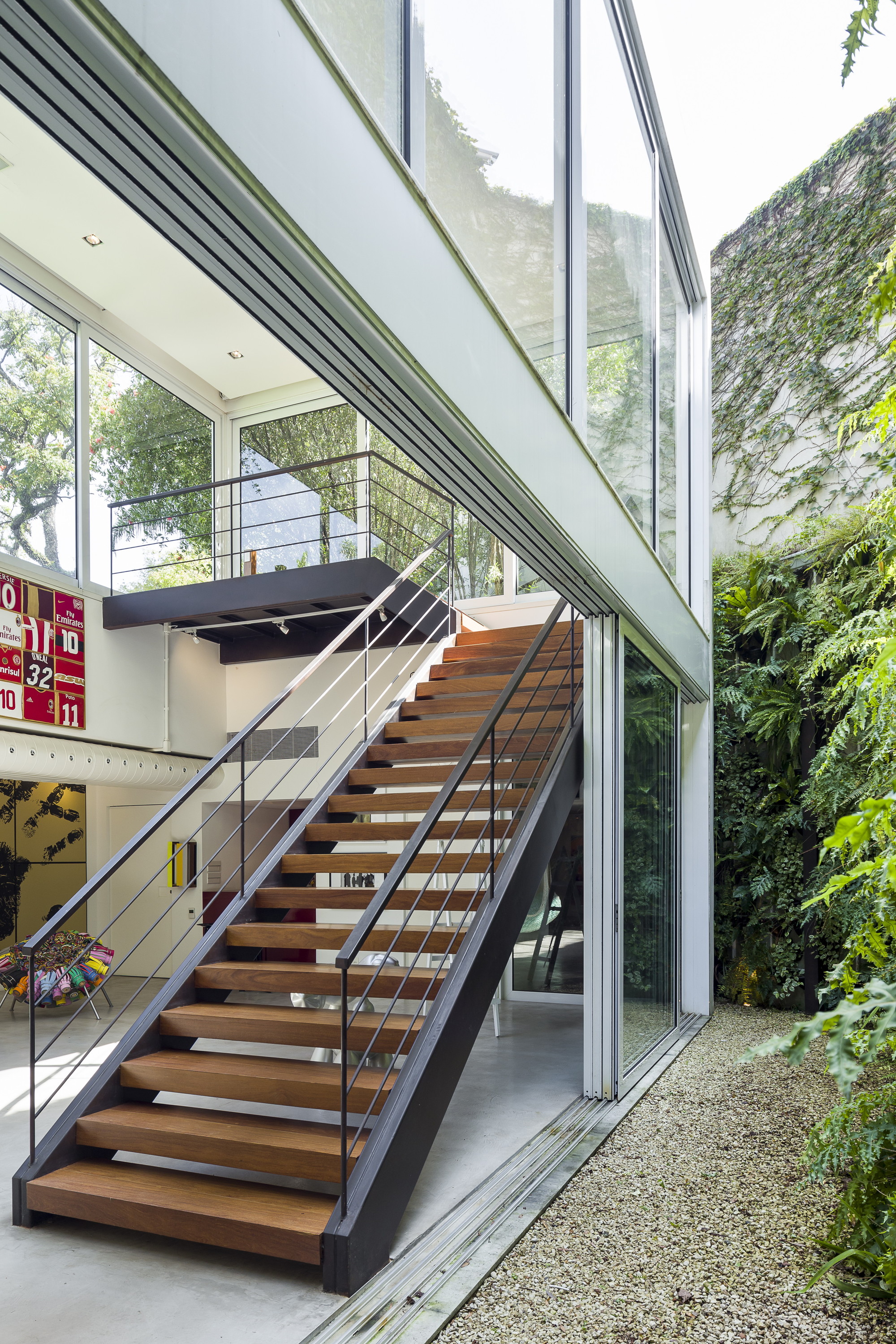 Steel and timber stair rising beside a glazed double-height wall, with a fern-covered wall and gravel path outside in daylight
