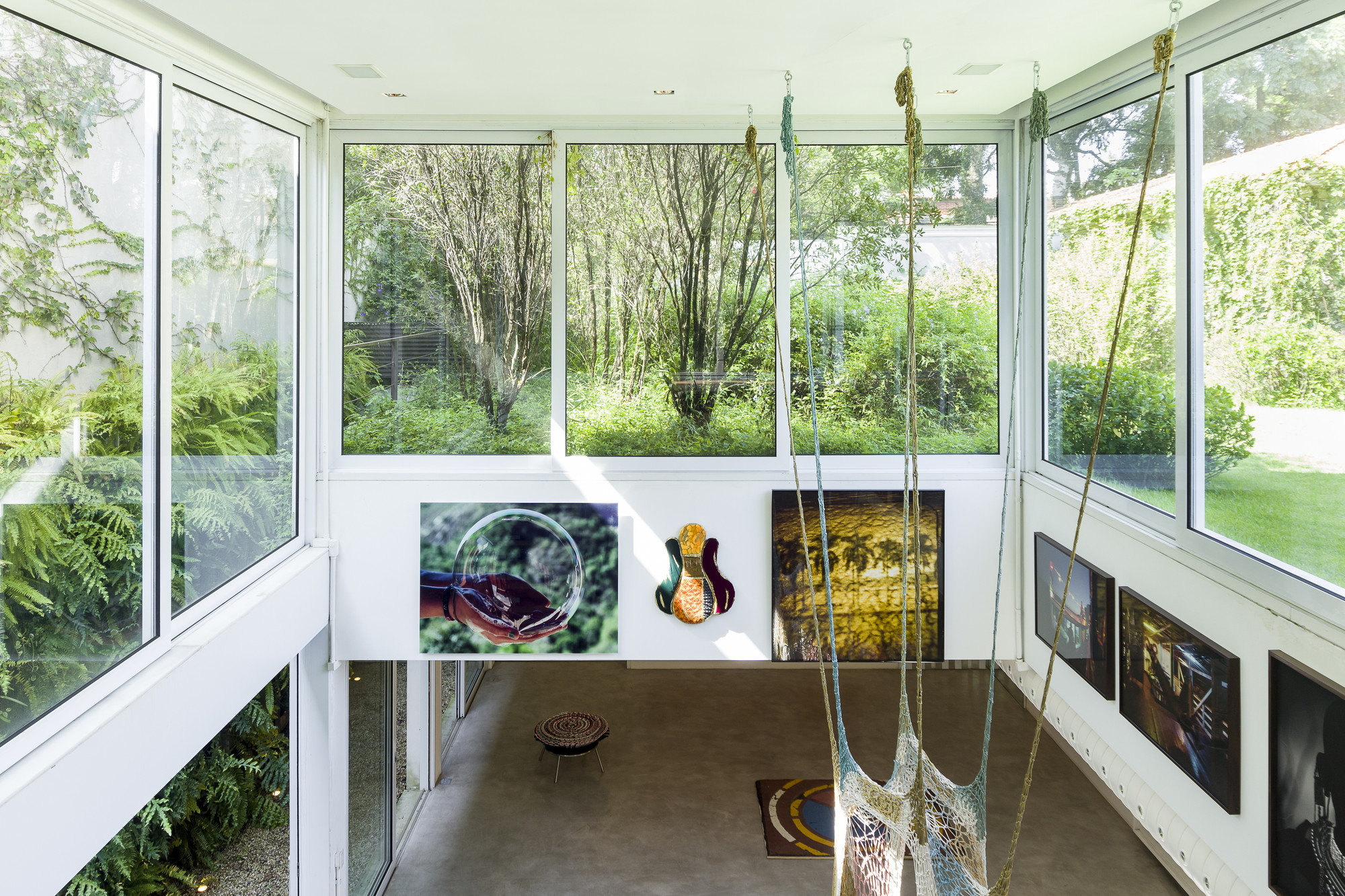 Double-height gallery room with framed photographs, a low stool and hanging woven ropes, with treetops visible through tall windows