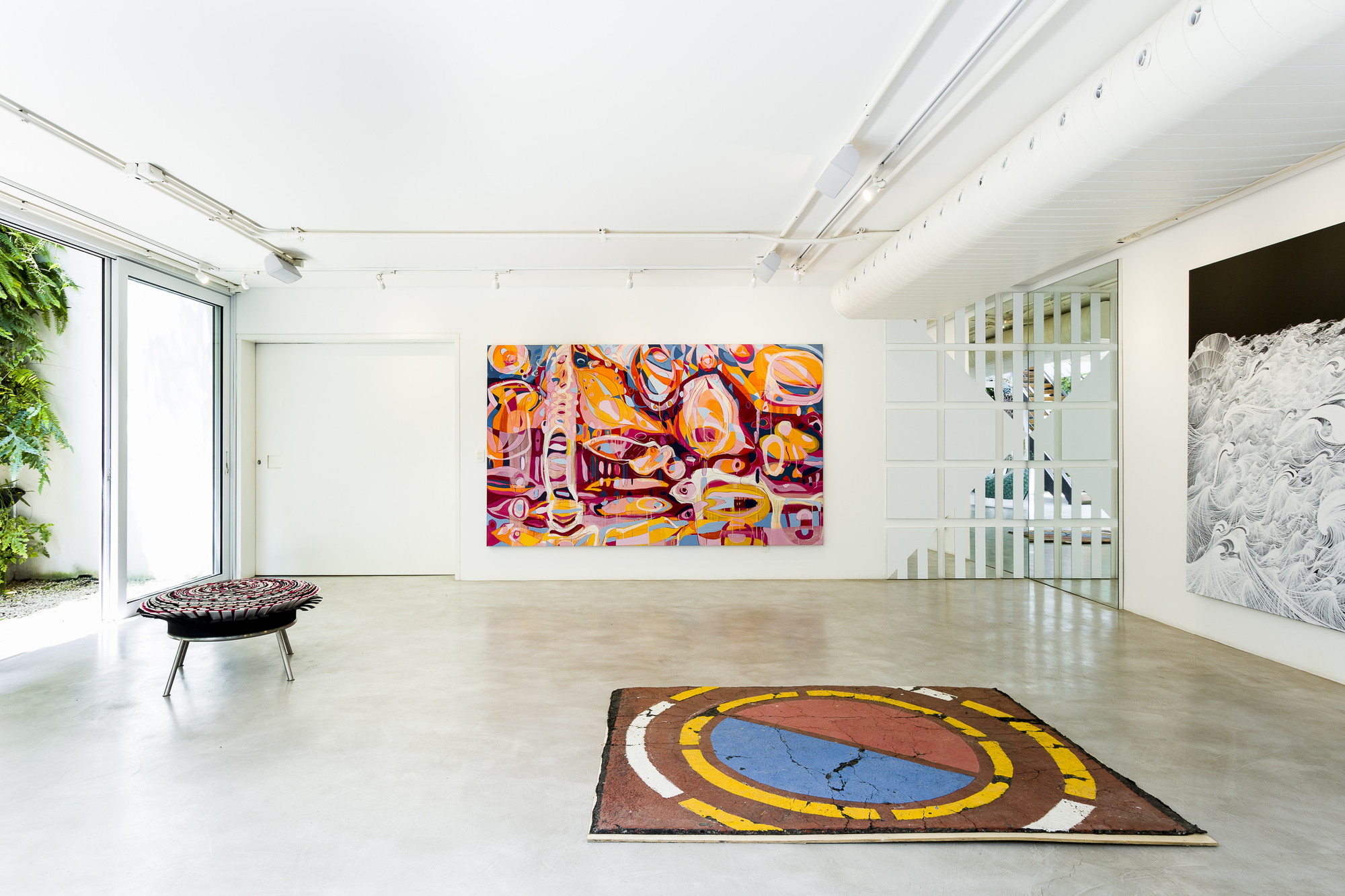 Gallery floor with a round painted rug and a woven stool before a large abstract canvas, daylight entering through glass on the left