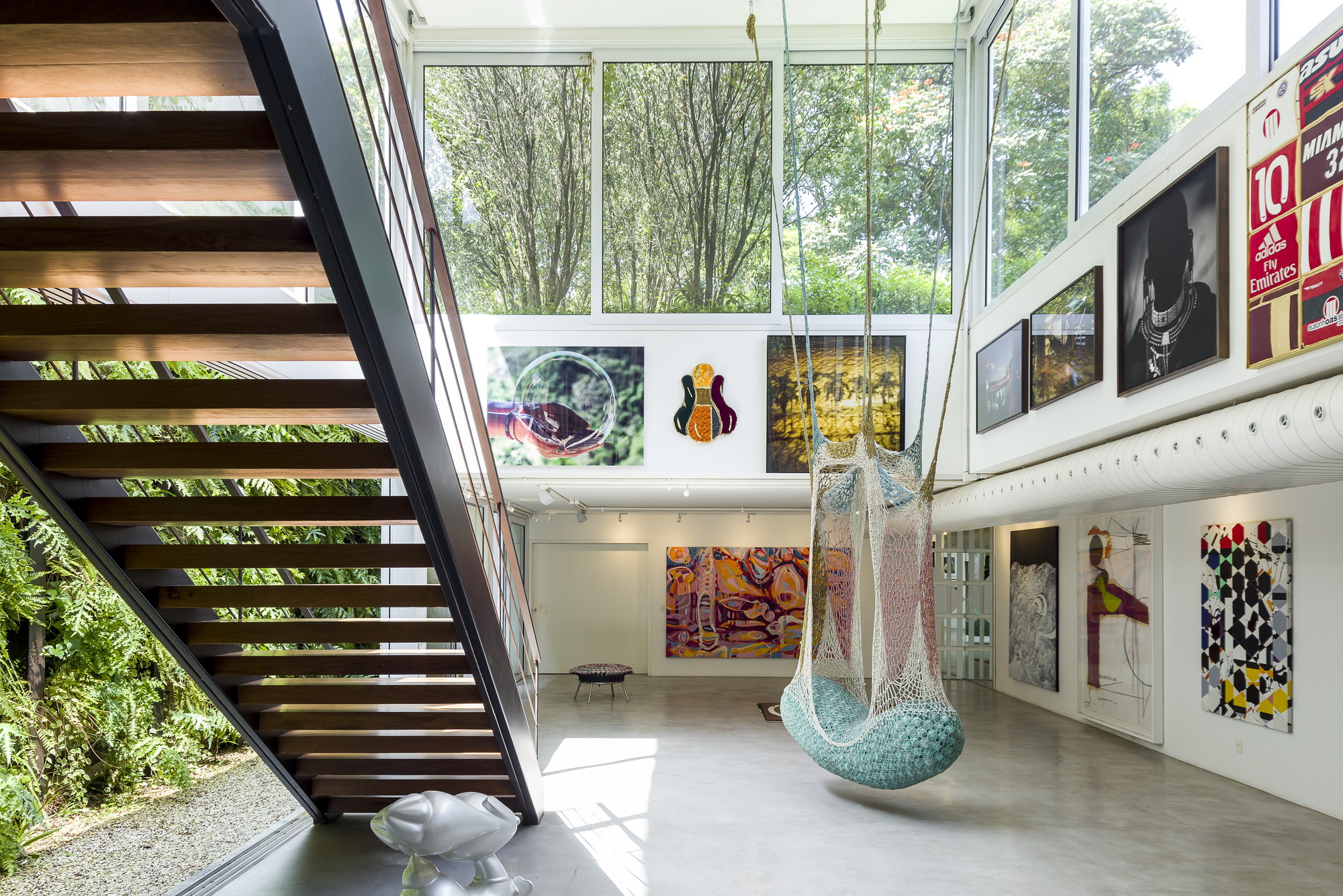 Double-height gallery with a timber-tread staircase, a hanging net chair and salon-style framed works beneath tall windows filled with treetops