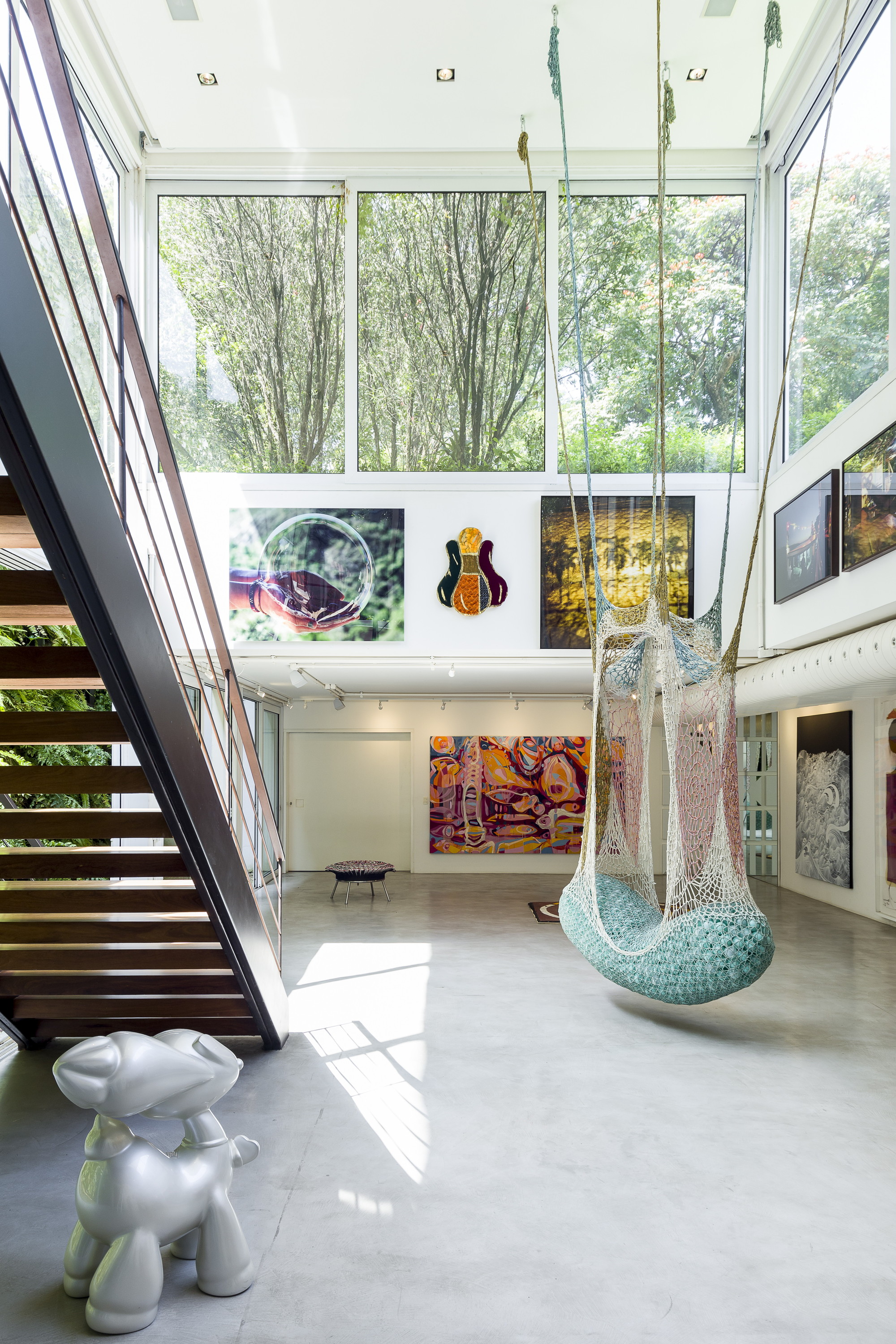 Vertical view of the double-height hall, with a silver animal sculpture, steel staircase and hanging net seat in a patch of sunlight