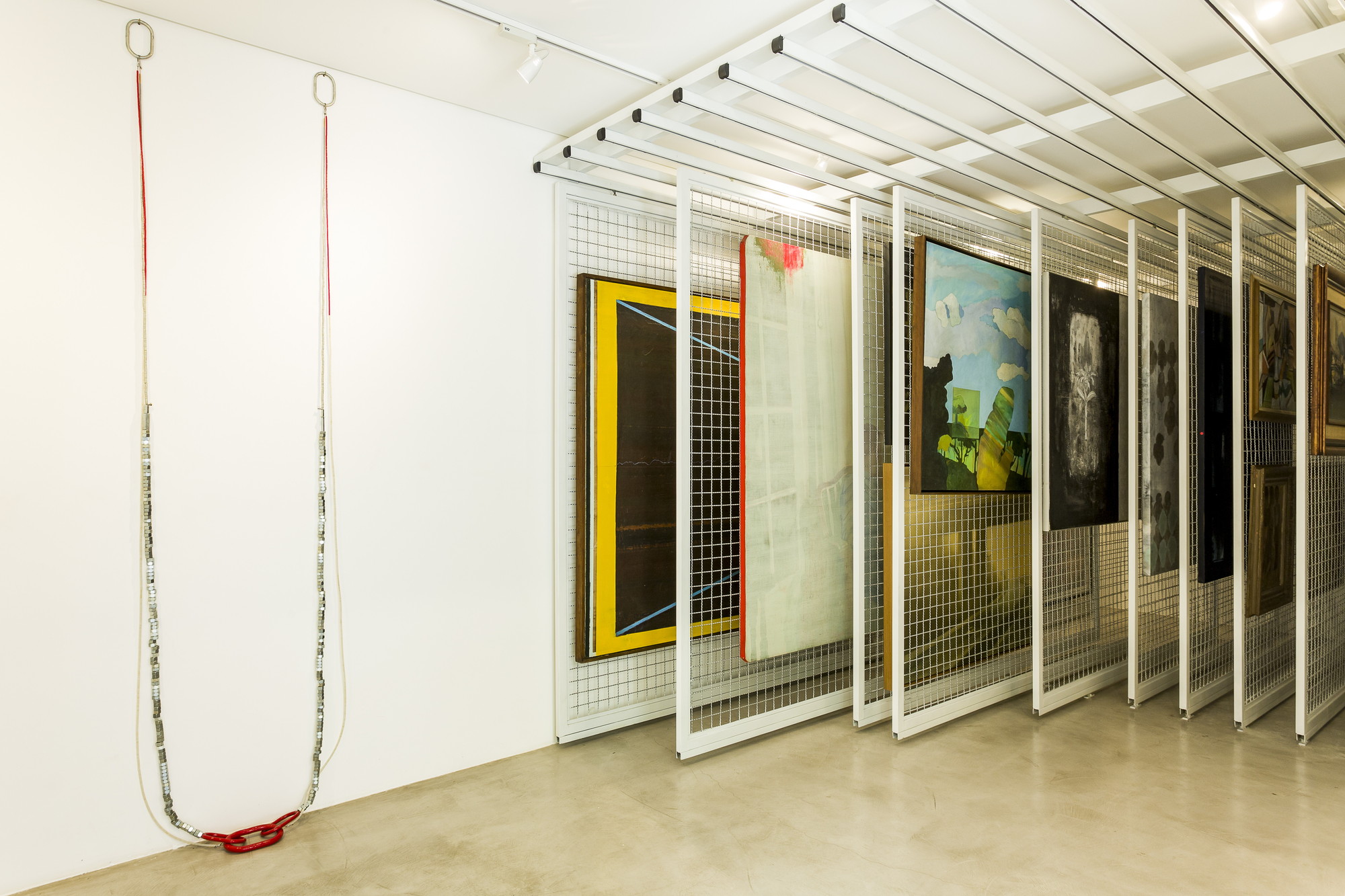 Sliding mesh racks holding framed paintings beside a white wall with a hanging rope and red chain piece
